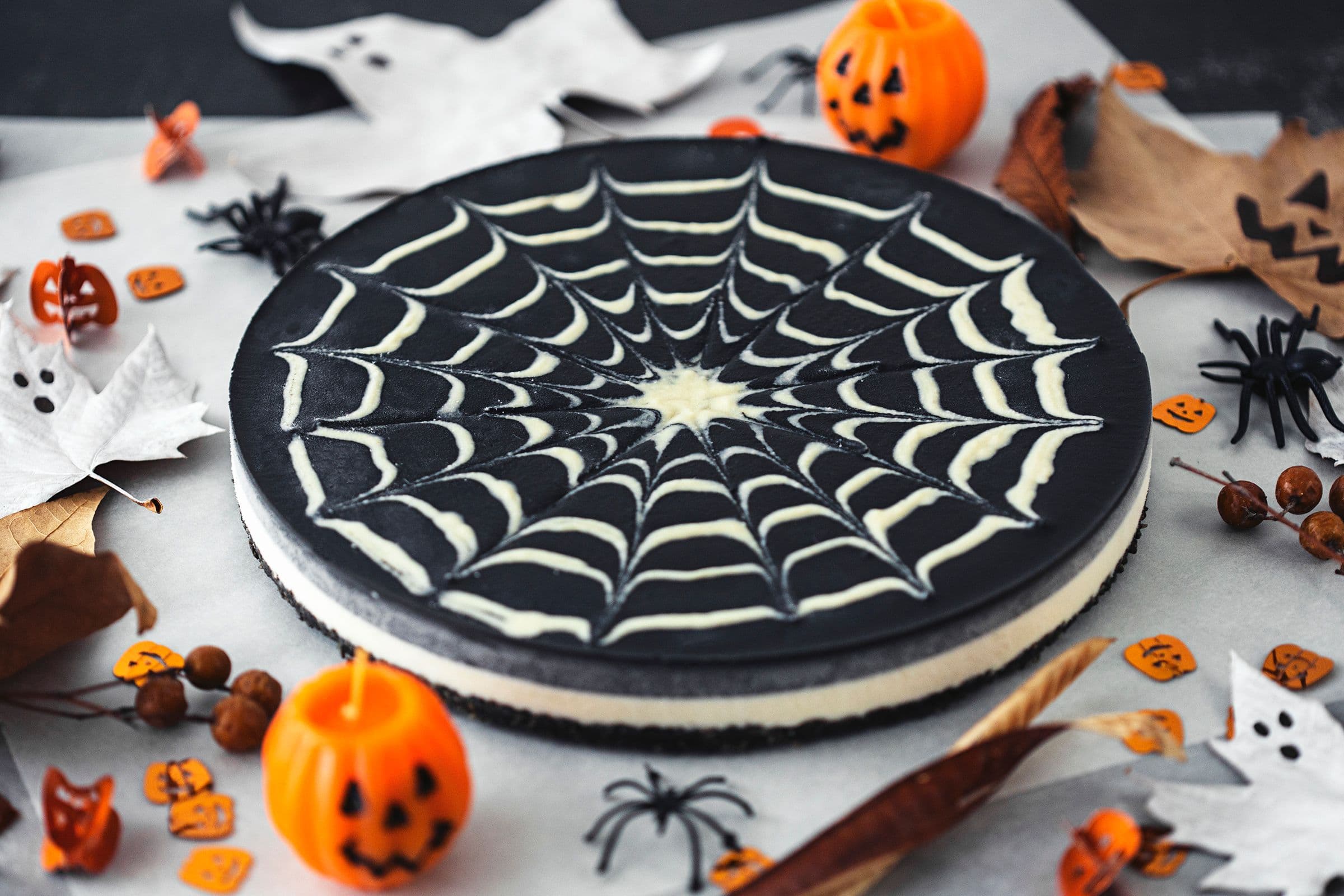 Cheesecake de chocolate duplo com padrão de teia de aranha, rodeado de abóboras, aranhas e decorações festivas.