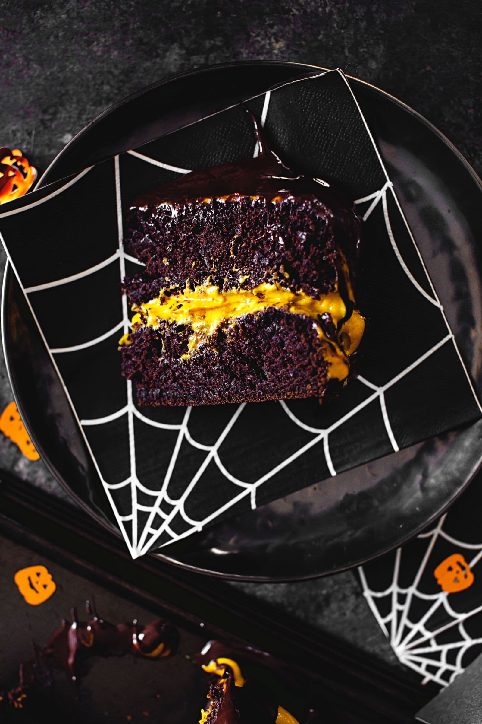 Slice of moist chocolate cake with bright orange cashew frosting, served on a black spiderweb-patterned napkin.