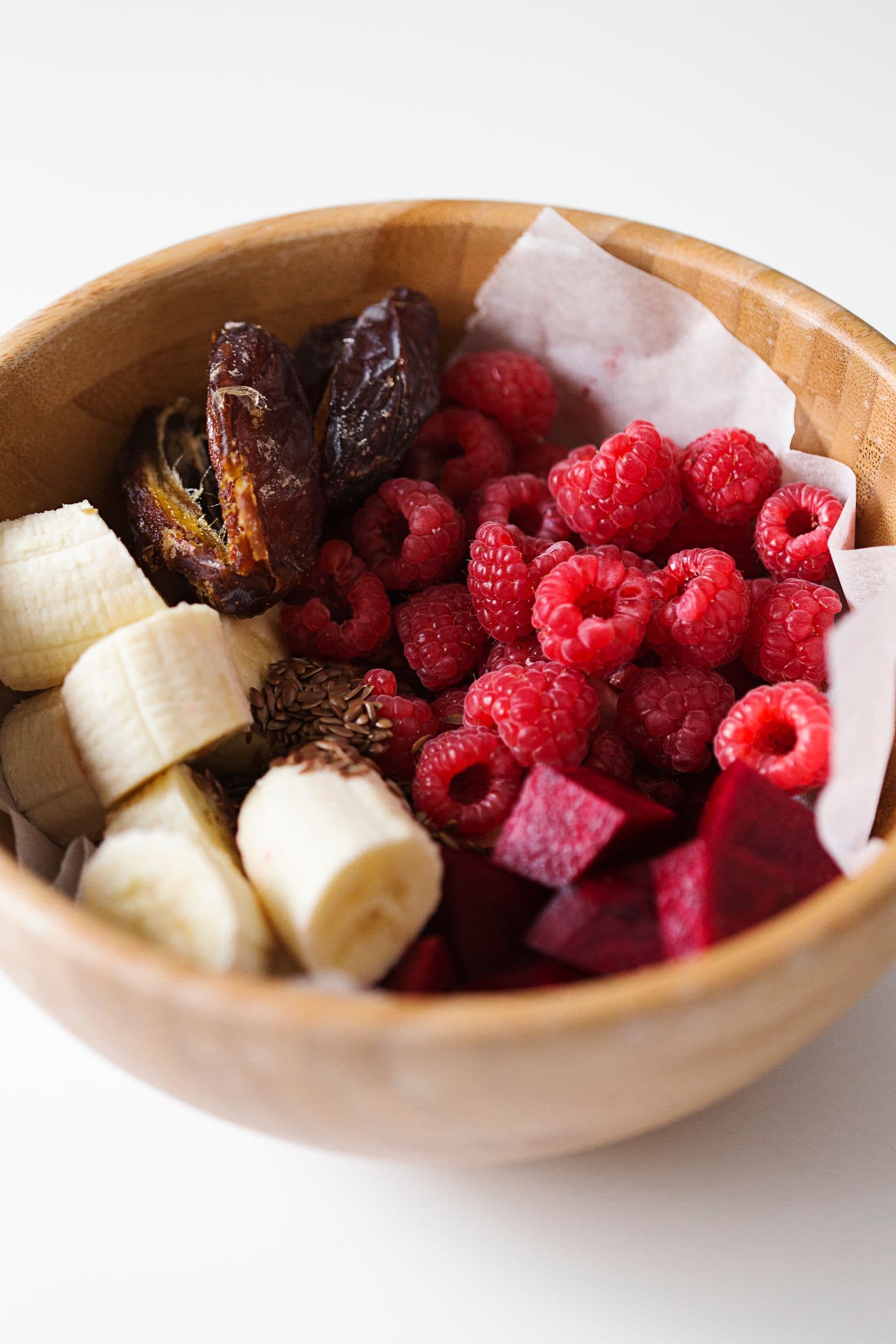 Taça de madeira com ingredientes para smoothie: banana, tâmaras, framboesas congeladas, beterraba e sementes de linhaça.