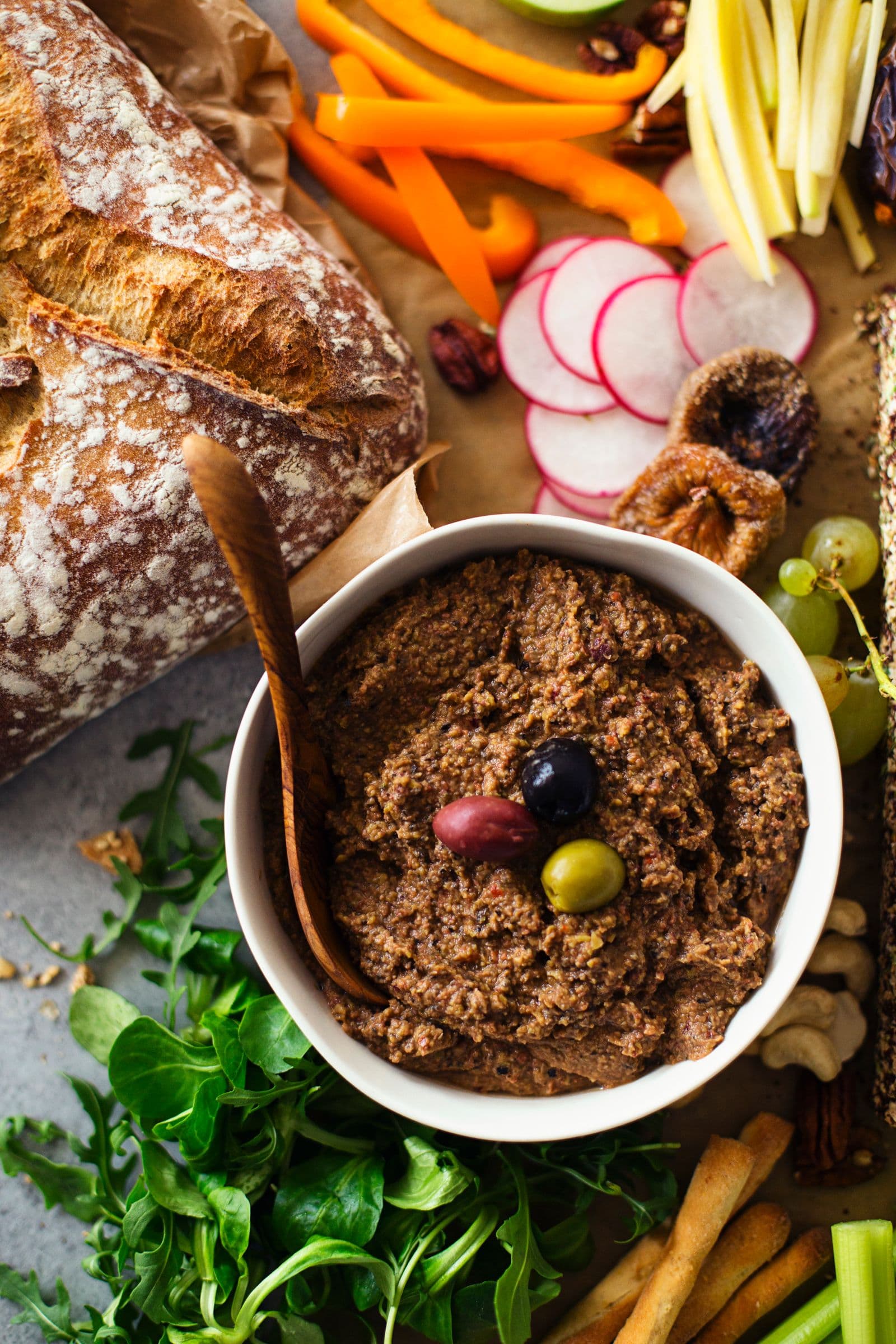 Small bowl of lentil and olive pâté garnished with olives, surrounded by fresh greens.