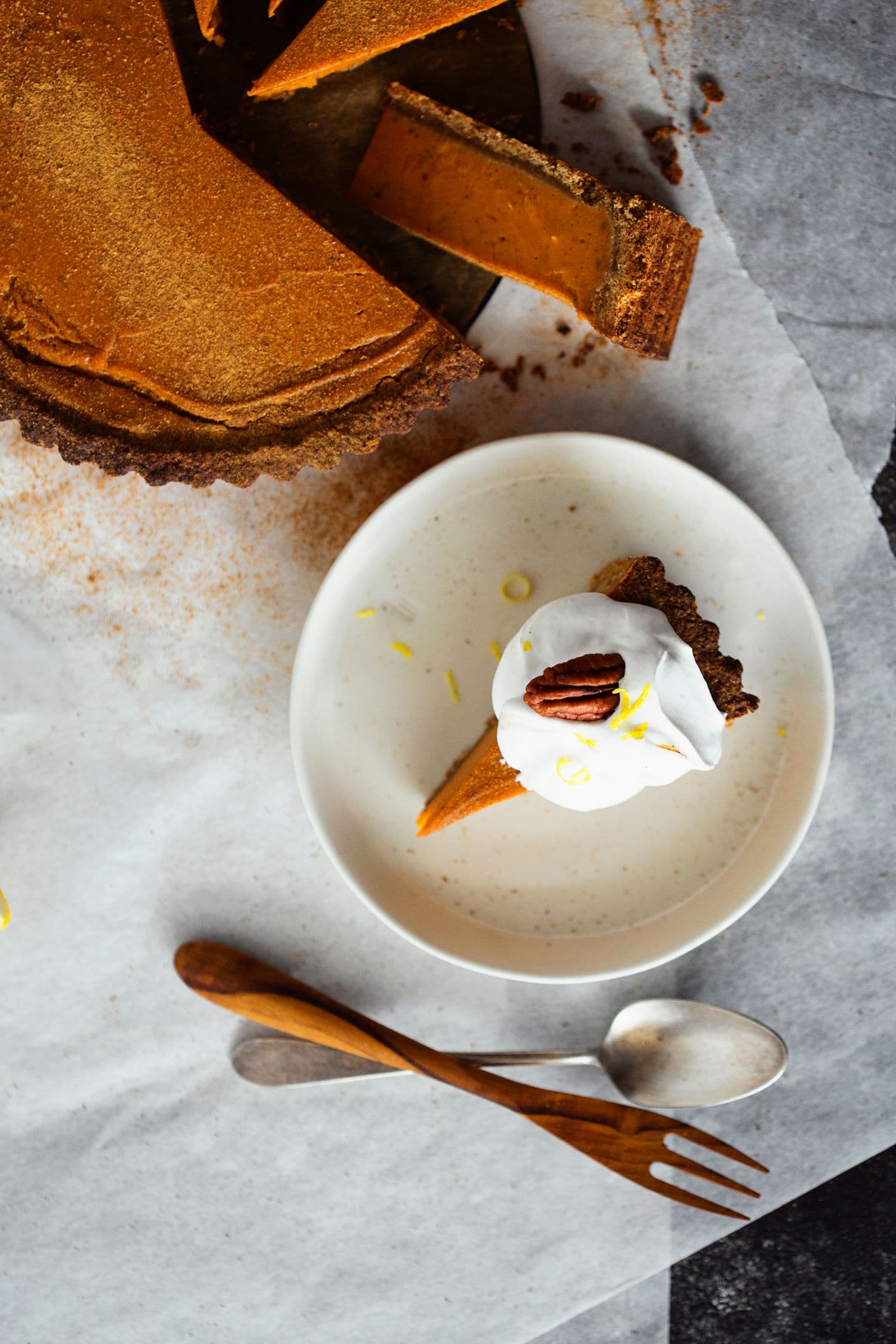 Slice of sweet potato pie topped with vegan whipped cream and a pecan, served on a white plate with a fork and spoon.