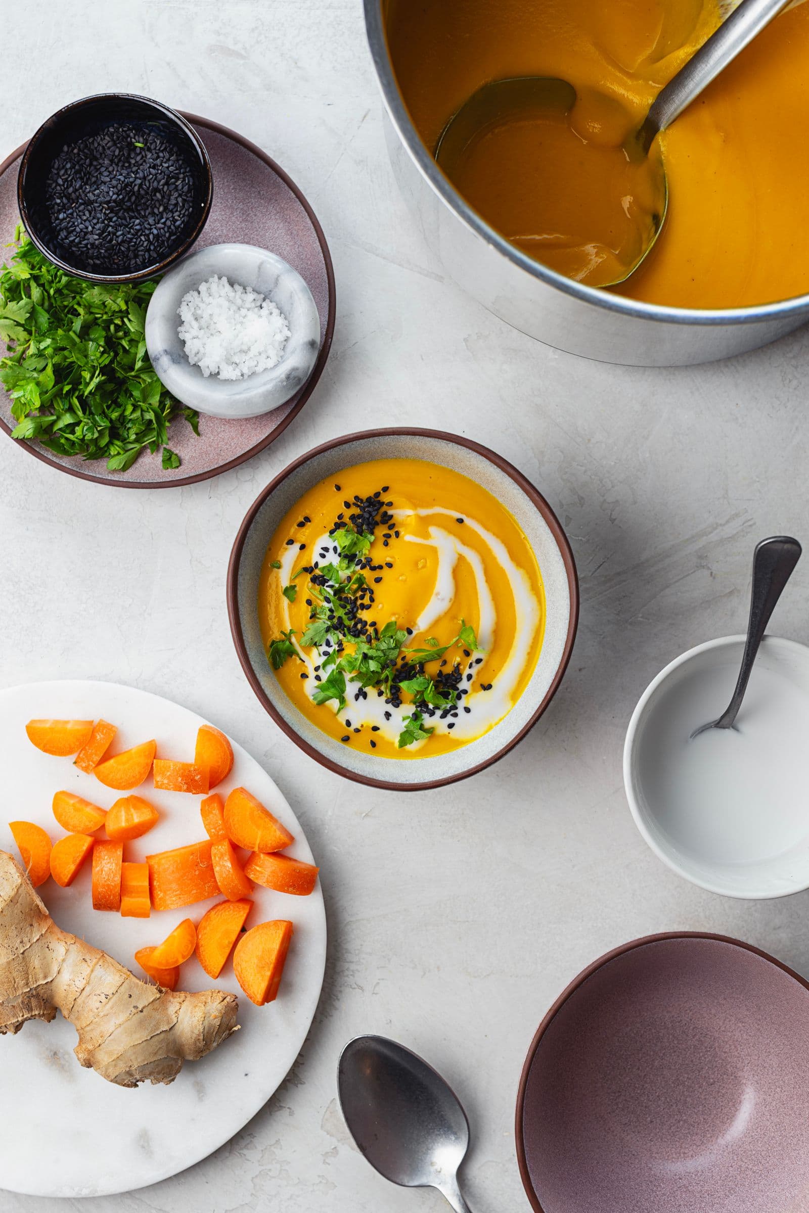 Bowl of creamy carrot soup topped with coconut milk, fresh parsley, and black seeds, with ingredients scattered around the table.