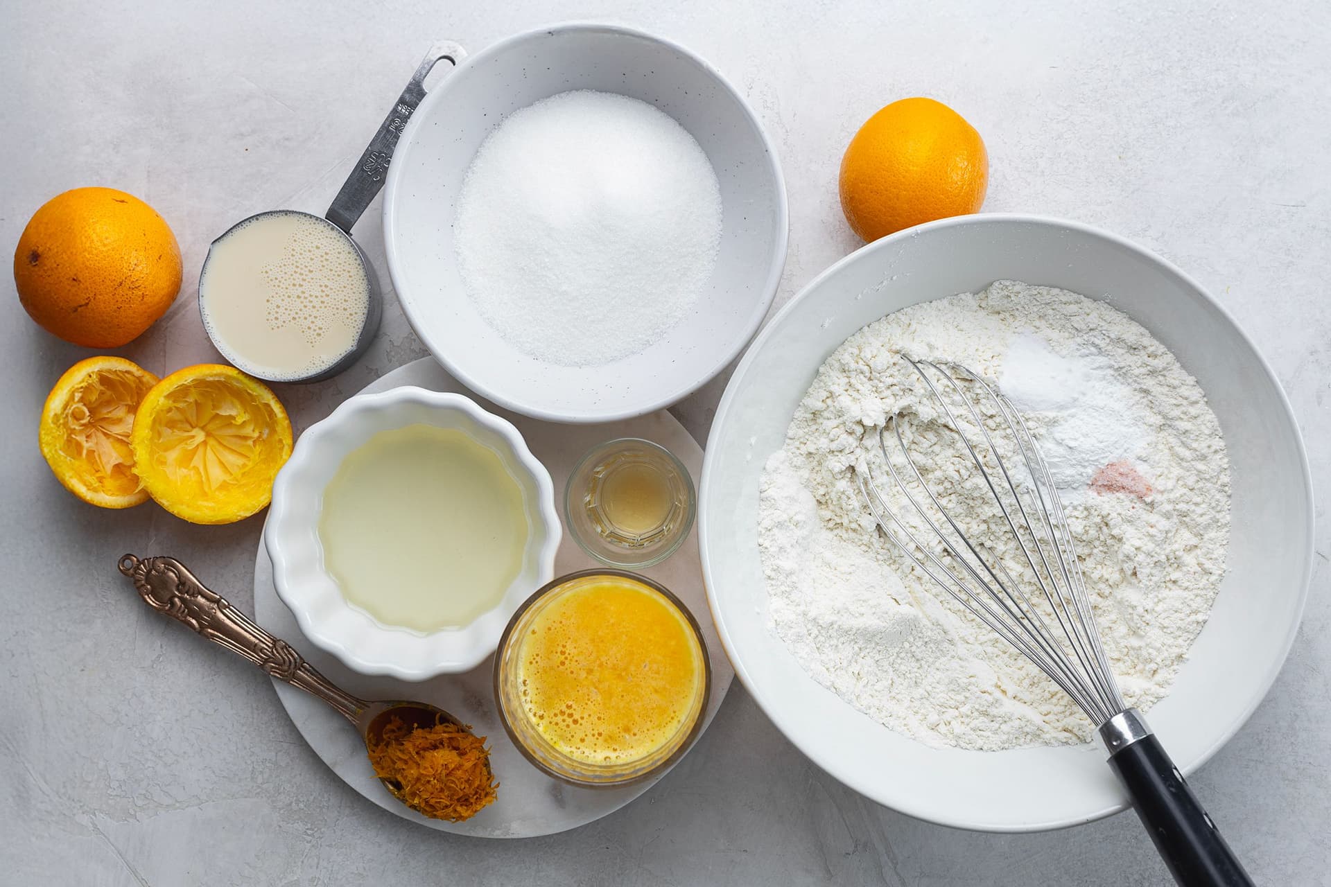 Ingredientes medidos para o bolo de laranja: farinha, açúcar, sumo e raspa de laranja, óleo e leite de soja, dispostos sobre uma superfície clara.