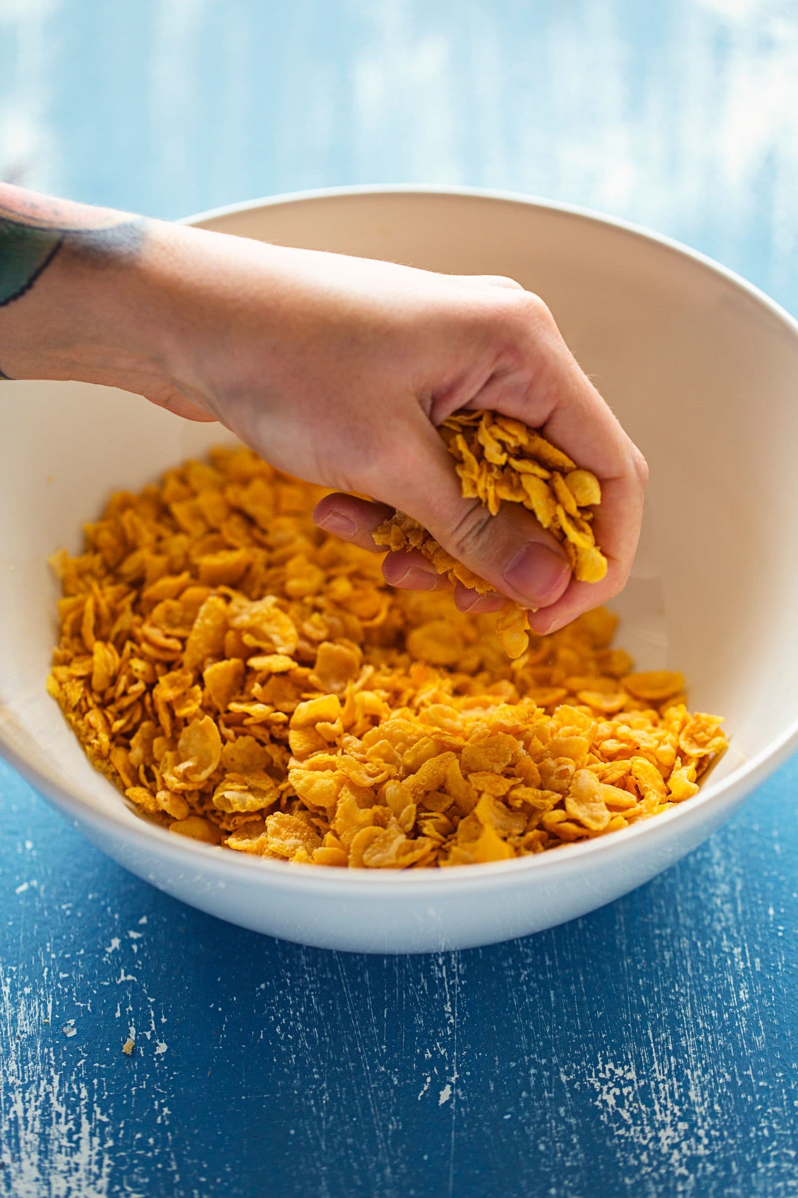 Mão a esmagar cornflakes para uma taça para preparar a base da granola.