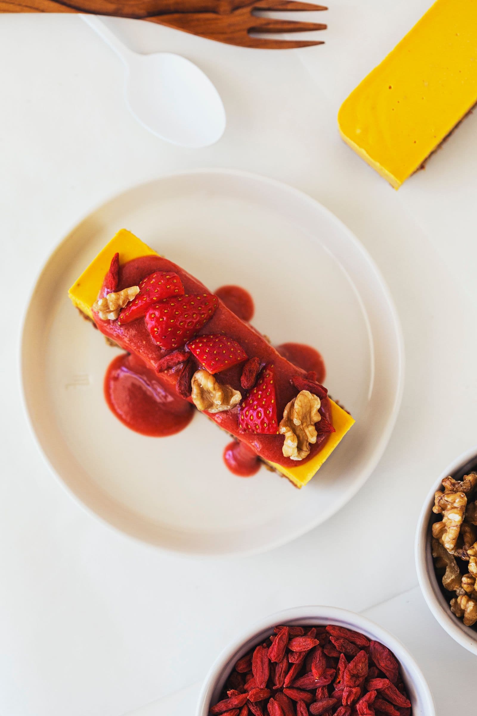 Uma fatia de cheesecake vegan de laranja e goji decorada com molho de morango, morangos frescos e nozes.