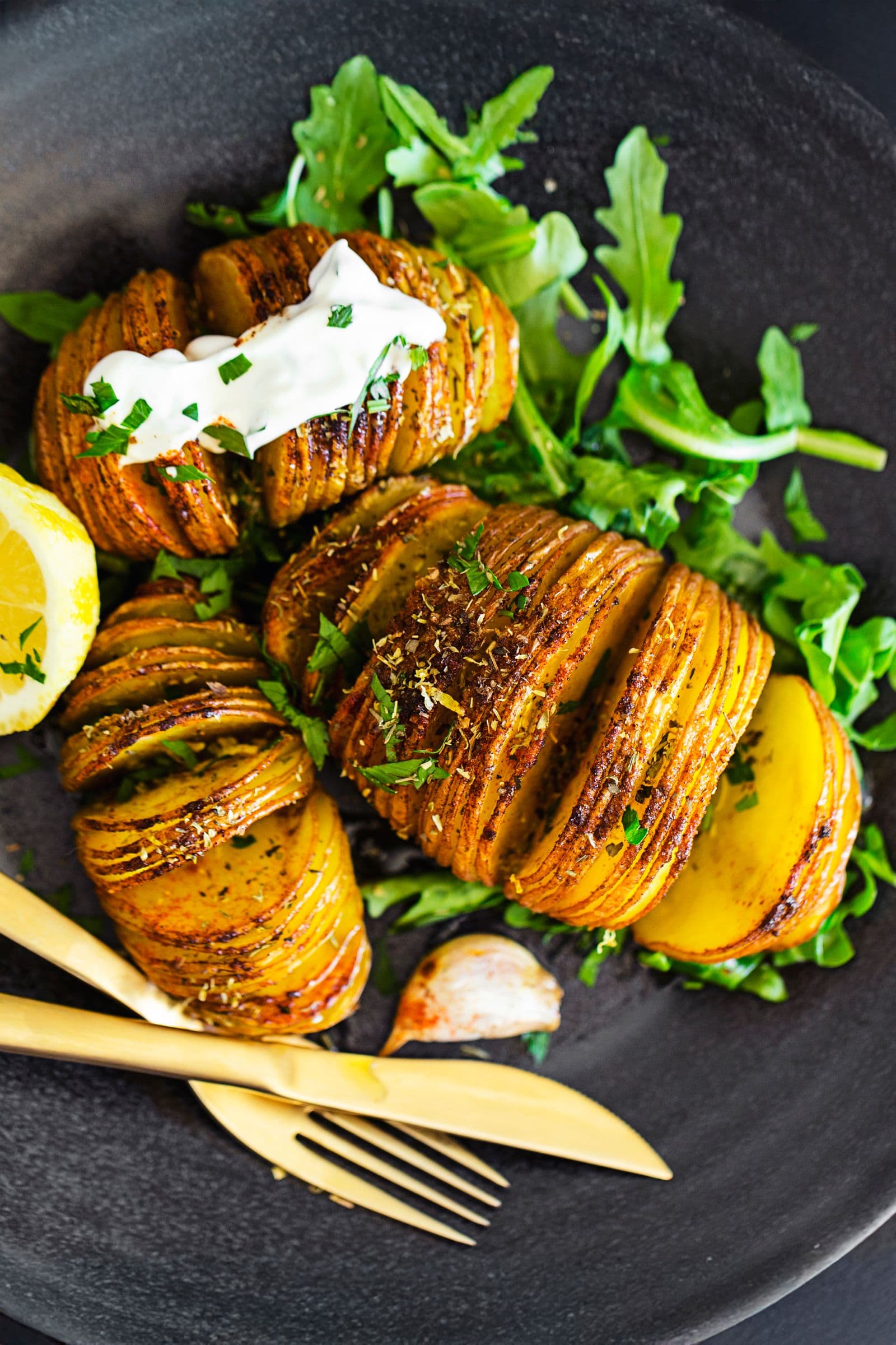 Golden Hasselback potatoes with herbs, spices and creamy sauce, served with fresh arugula on a black plate.