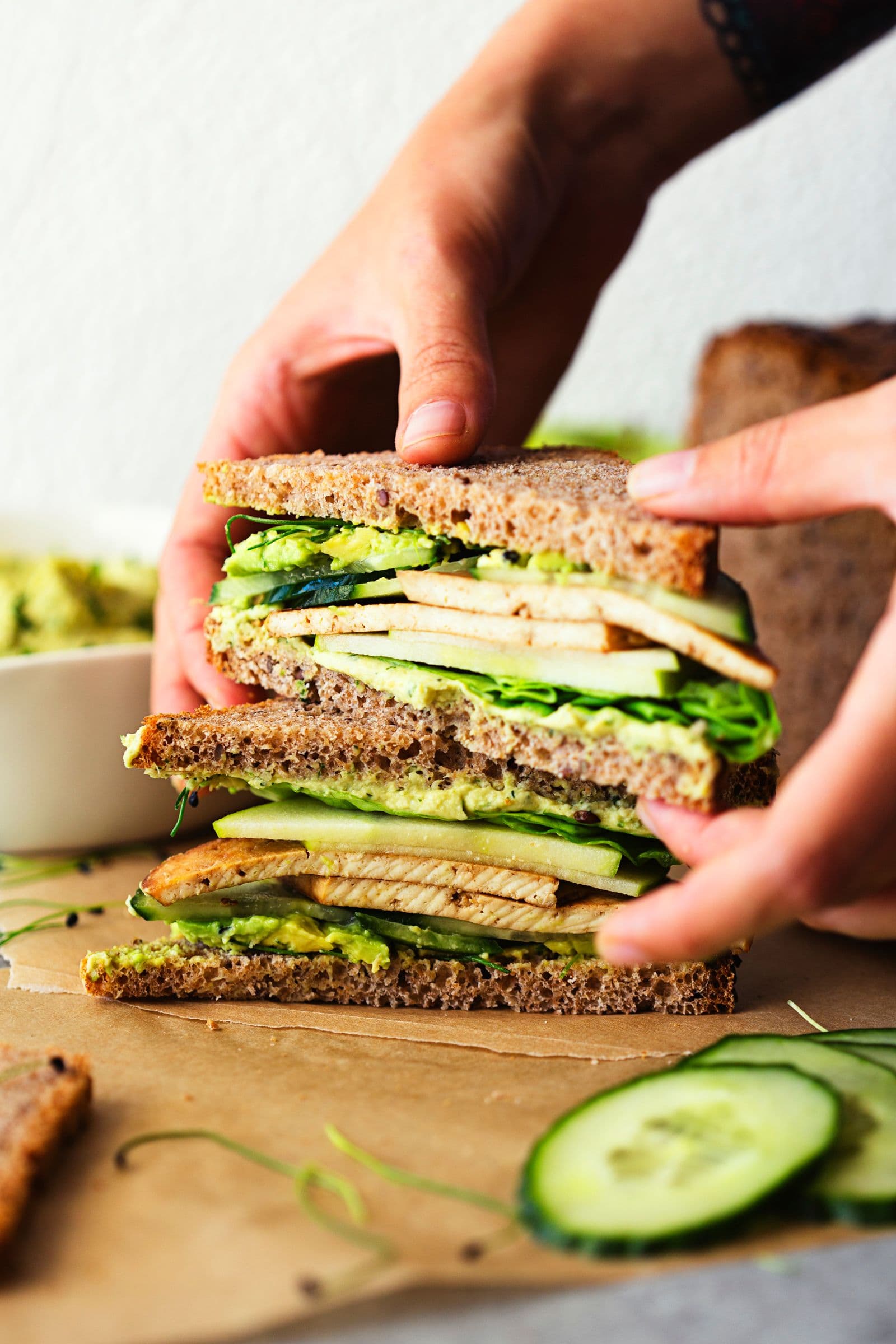 Close-up of hands holding stacked green tofu sandwich with cucumber slices.