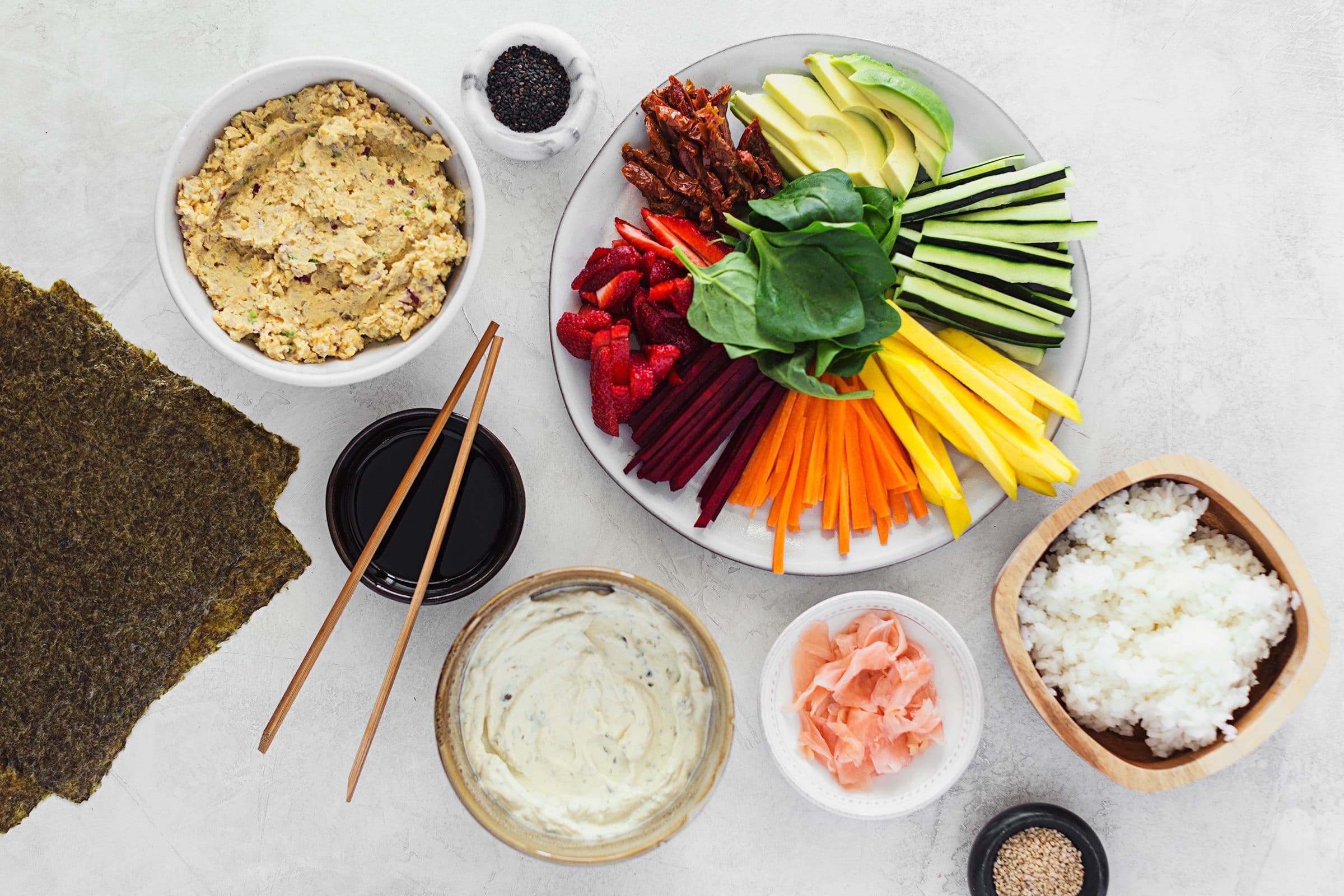 Ingredientes para sushi vegan dispostos numa mesa: algas nori, arroz, vegetais fatiados, pasta de tofu e molho para acompanhar.