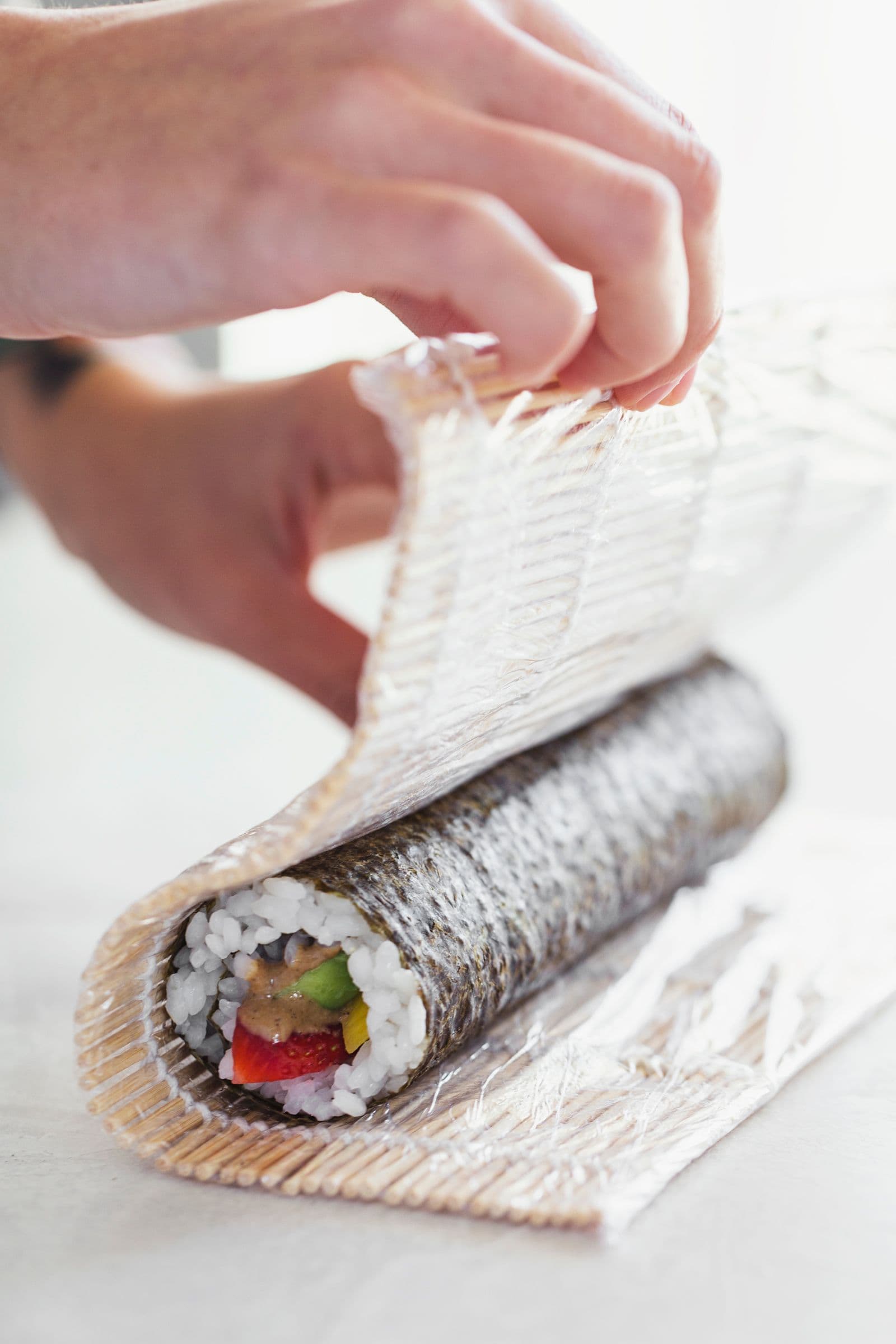 Mãos a enrolar sushi vegan com a ajuda de uma esteira de bambu.