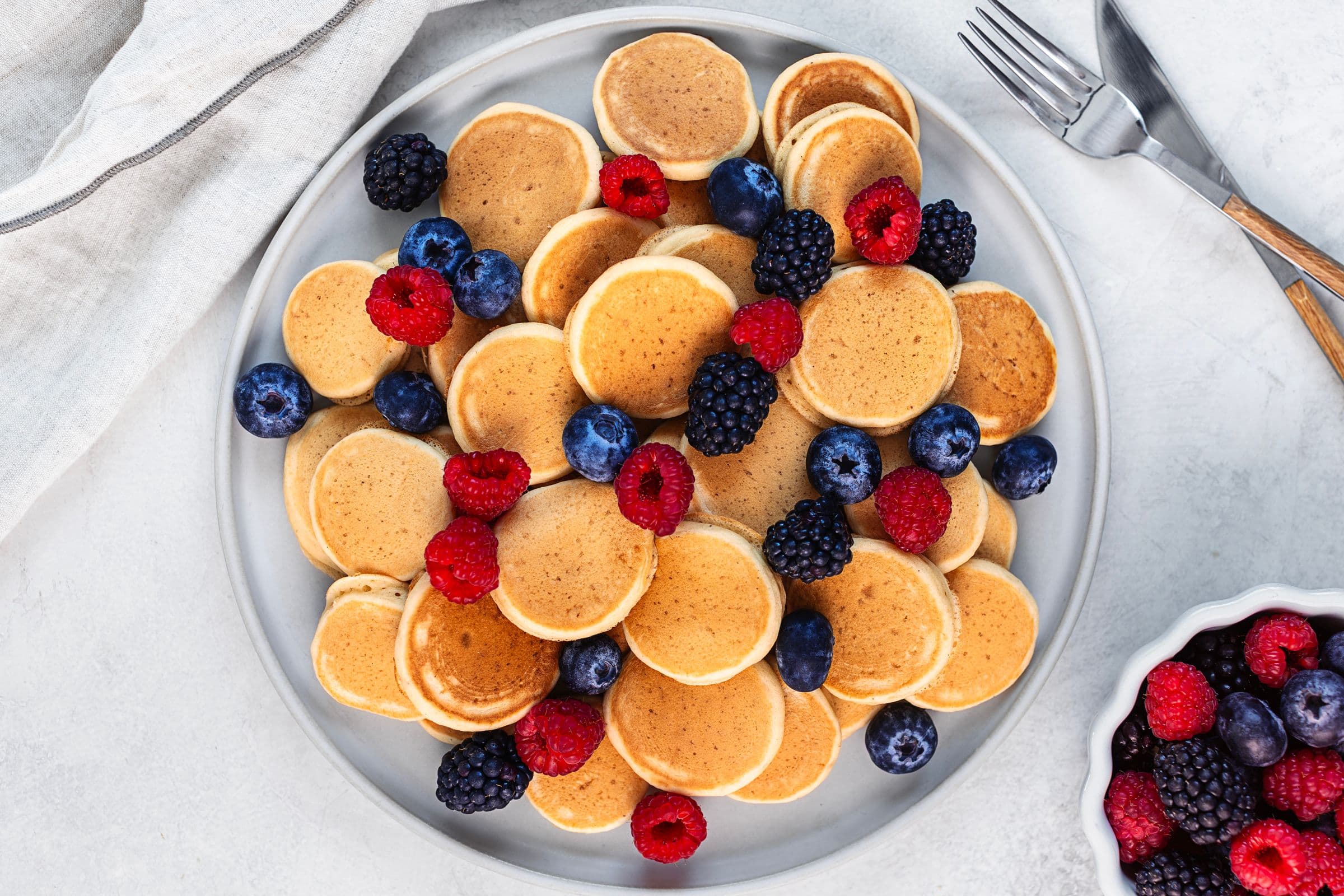 Prato de mini panquecas vegan com framboesas, mirtilos e amoras frescas por cima, com talheres ao lado