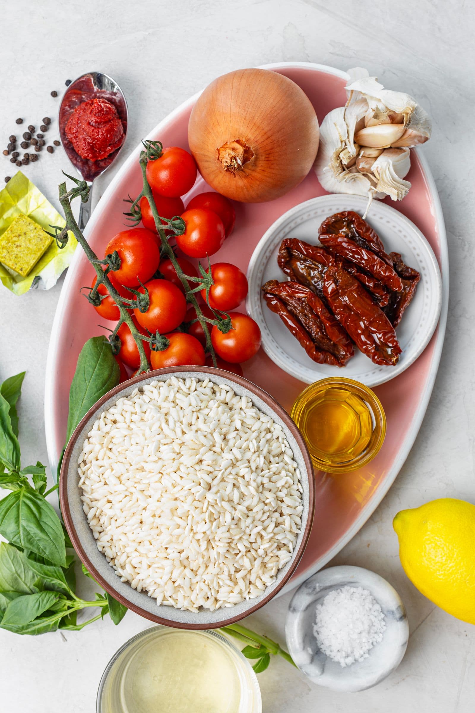 Overhead view of tomato risotto ingredients: cherry tomatoes on the vine, onion, garlic, sun-dried tomatoes, risotto rice, olive oil, tomato paste, stock cubes and fresh basil.