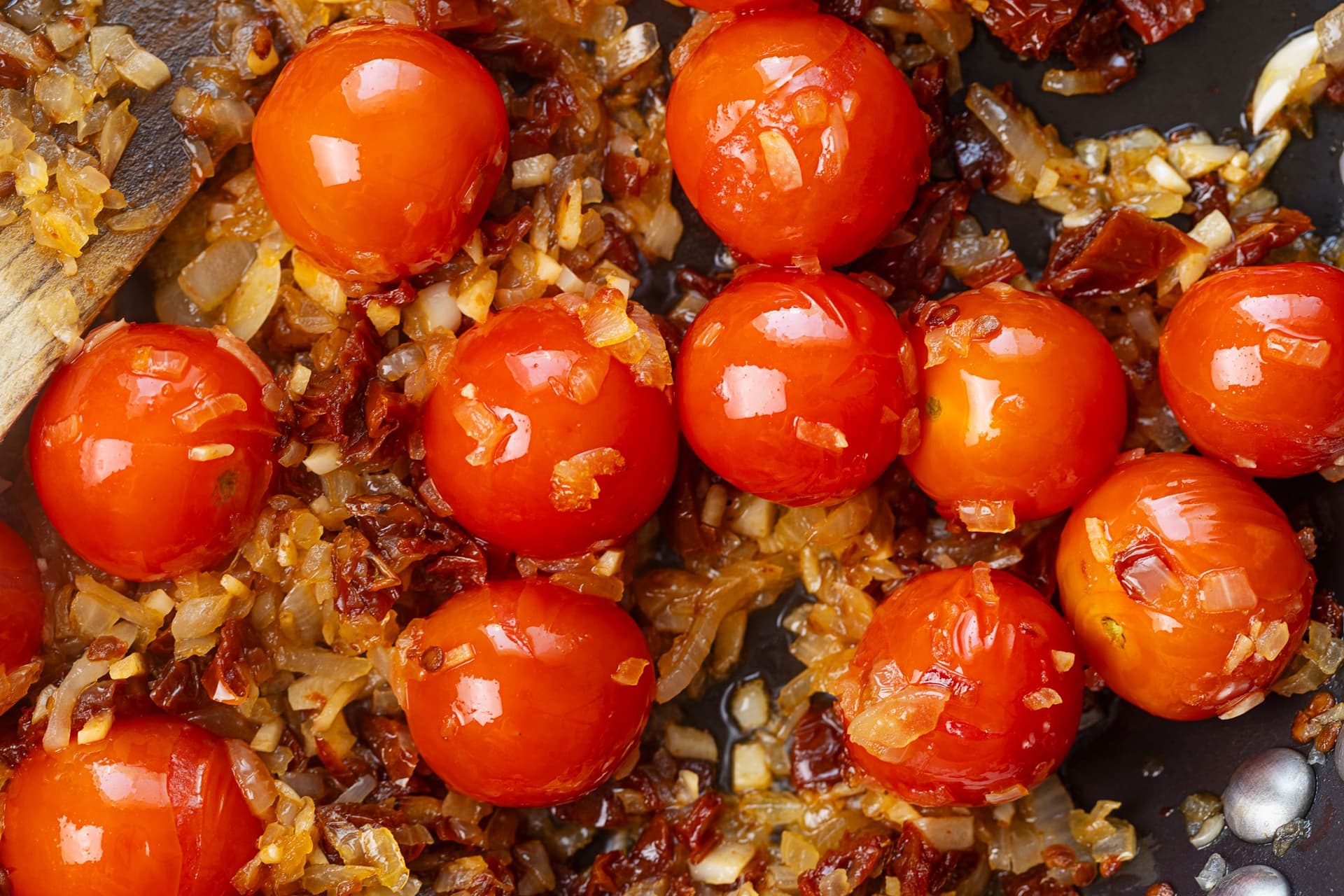 Cherry tomatoes sizzling with chopped onion, garlic and sun-dried tomatoes in a pan.