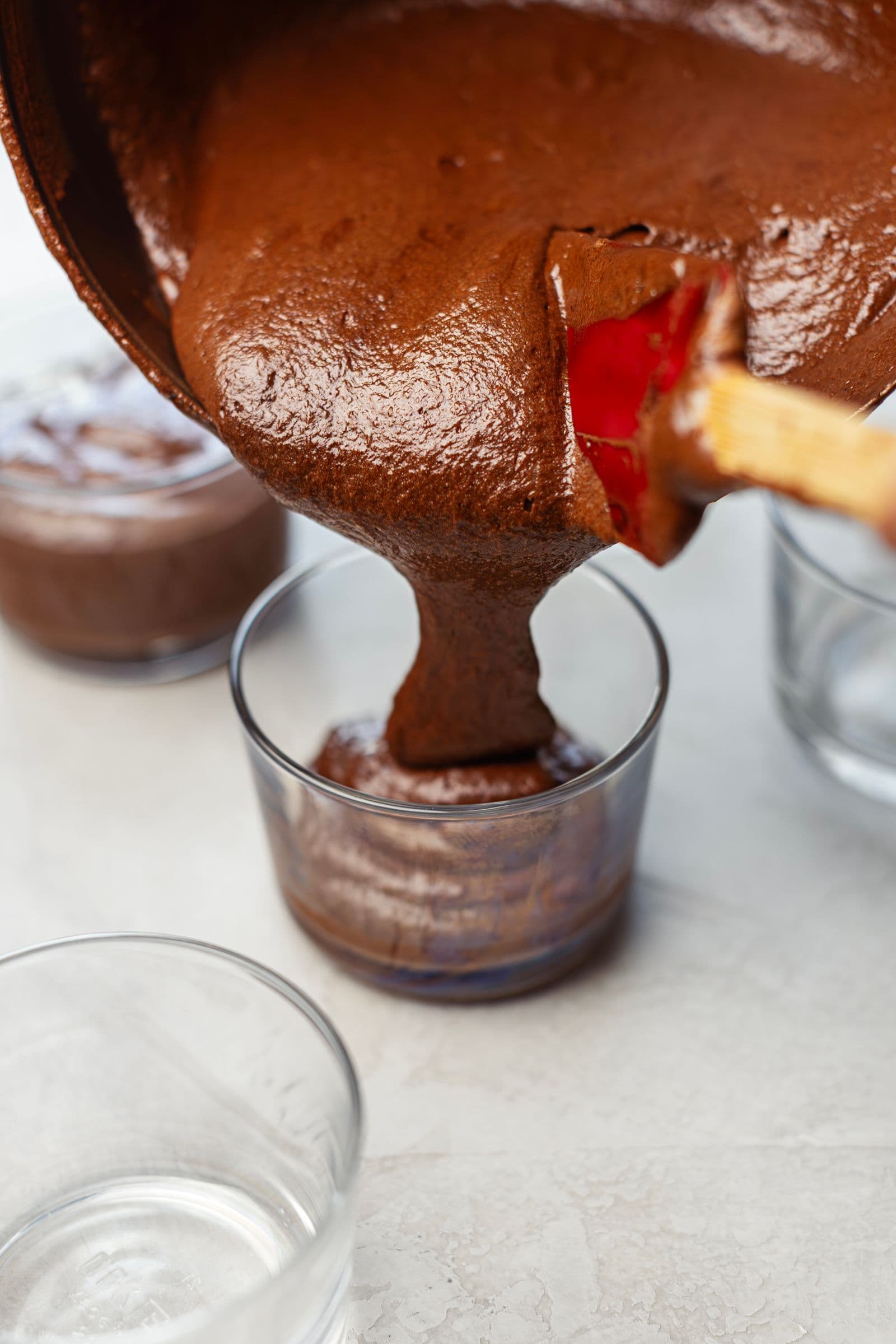 Vegan chocolate mousse being spooned into individual glass cups with a spatula.