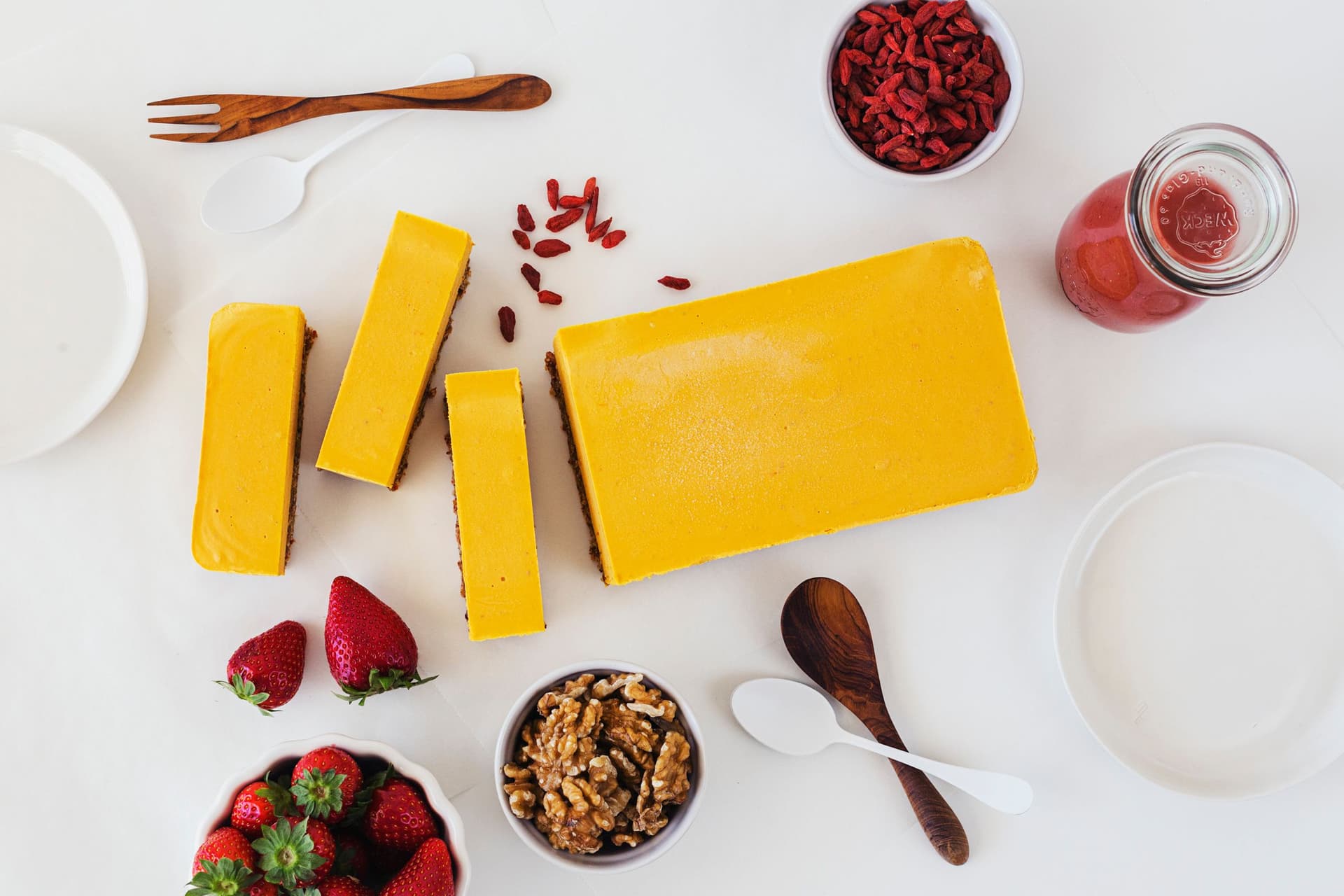 Barra de cheesecake laranja sobre mesa branca, com bagas goji e morangos espalhados em volta.