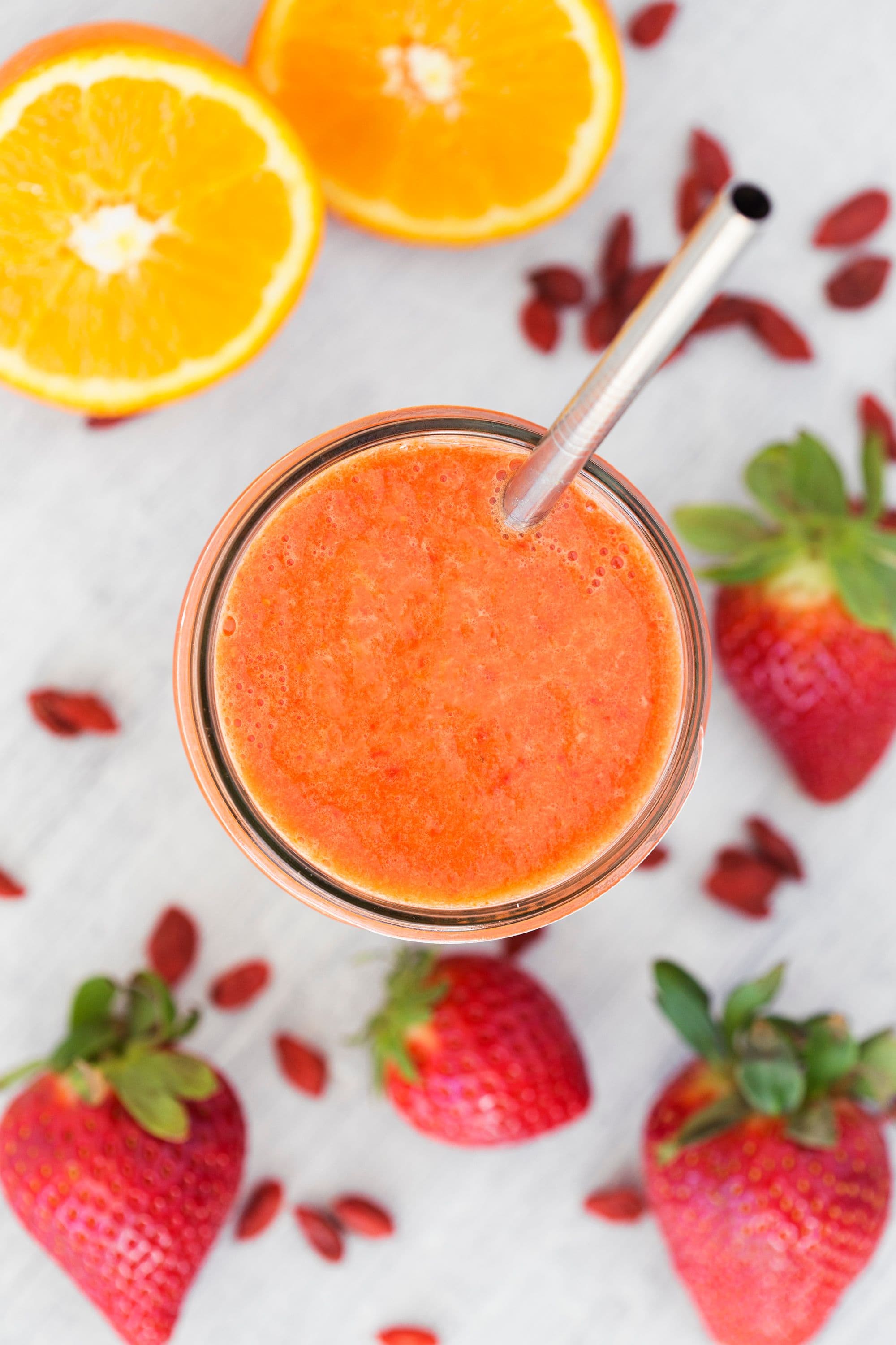Copo com smoothie visto de cima, rodeado por morangos, laranja e bagas goji.
