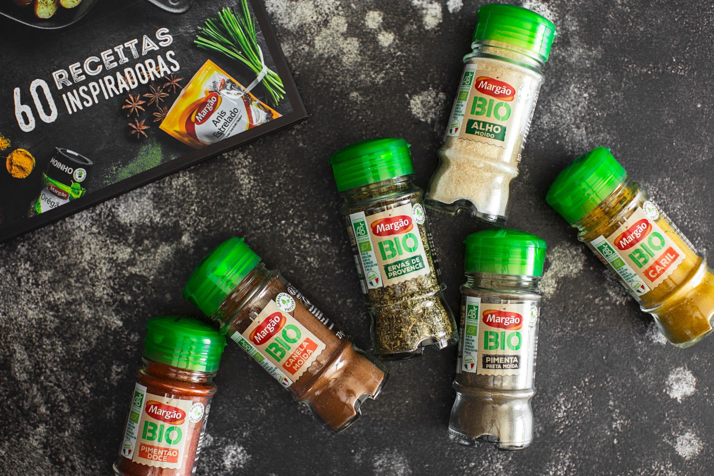 Set of Margão Bio spices, including garlic powder, curry, sweet paprika, cinnamon, black pepper, and herbes de Provence, on a textured black background.