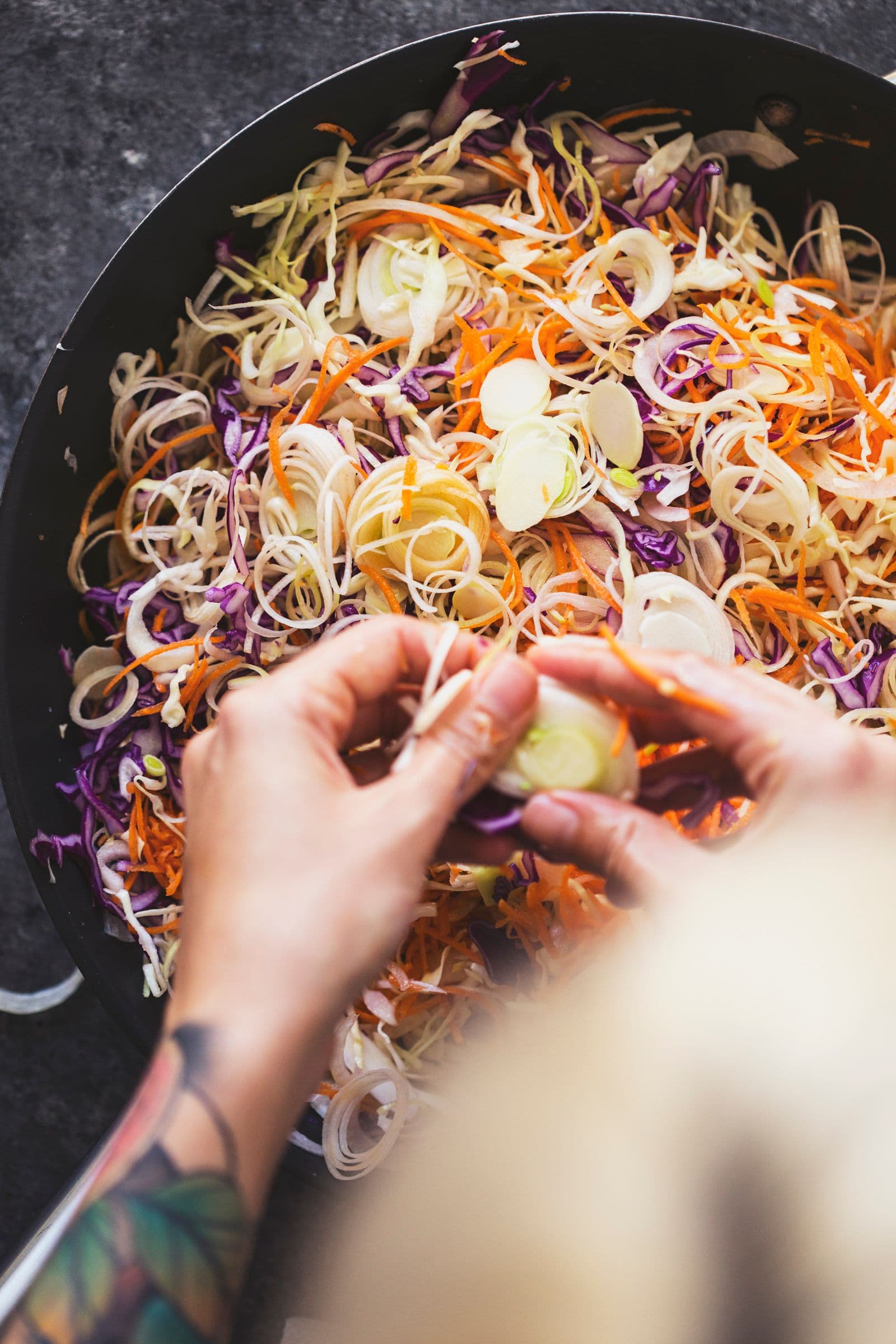 Hands stirring thinly sliced vegetables in a pan before sautéing.