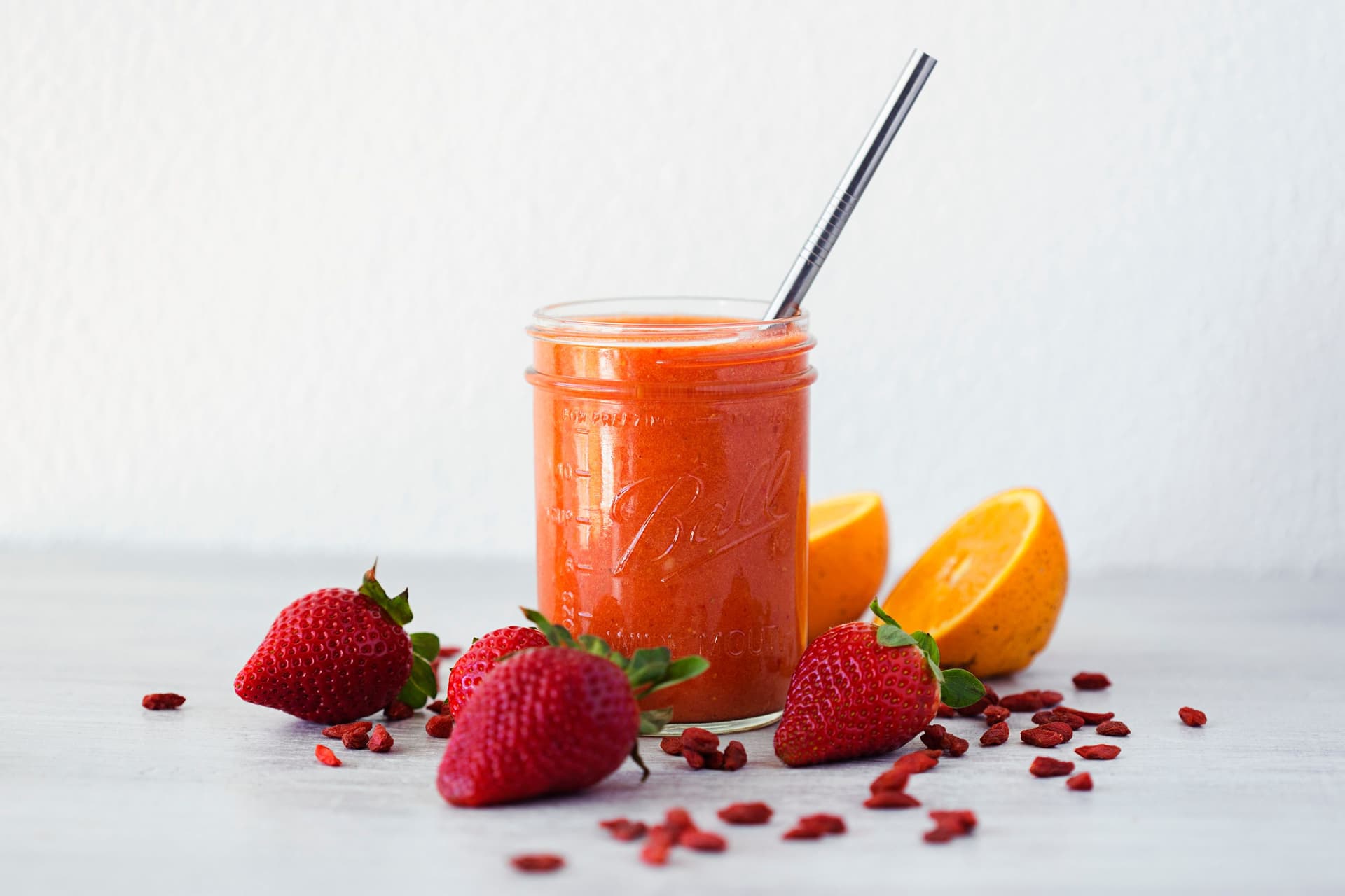 Copo com palhinha de smoothie de morango, laranja e bagas goji, com morangos frescos, gomos de laranja e bagas goji secas à volta.