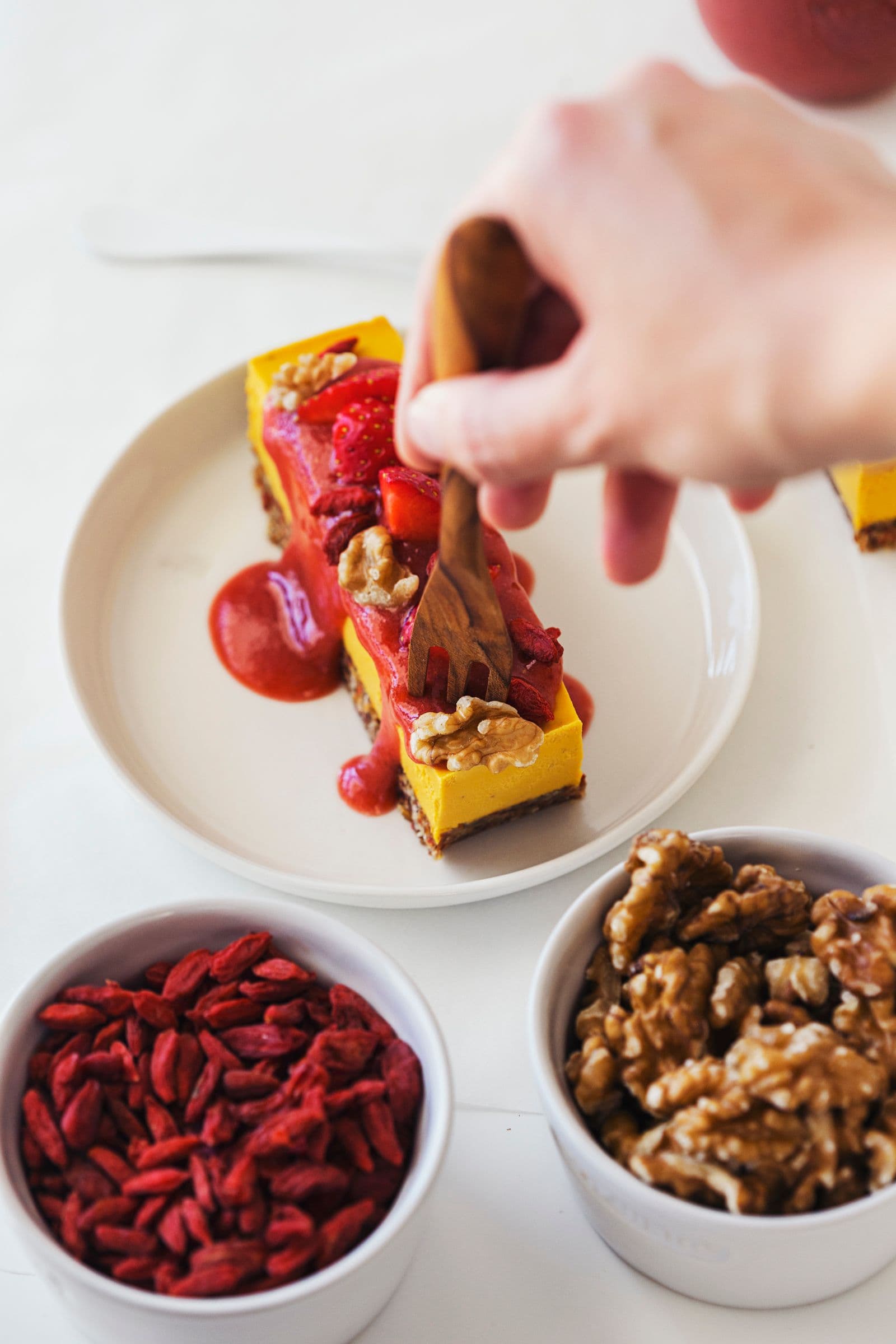 Hand holding wooden fork and digging into a slice of raw vegan cheesecake with fruity topping.