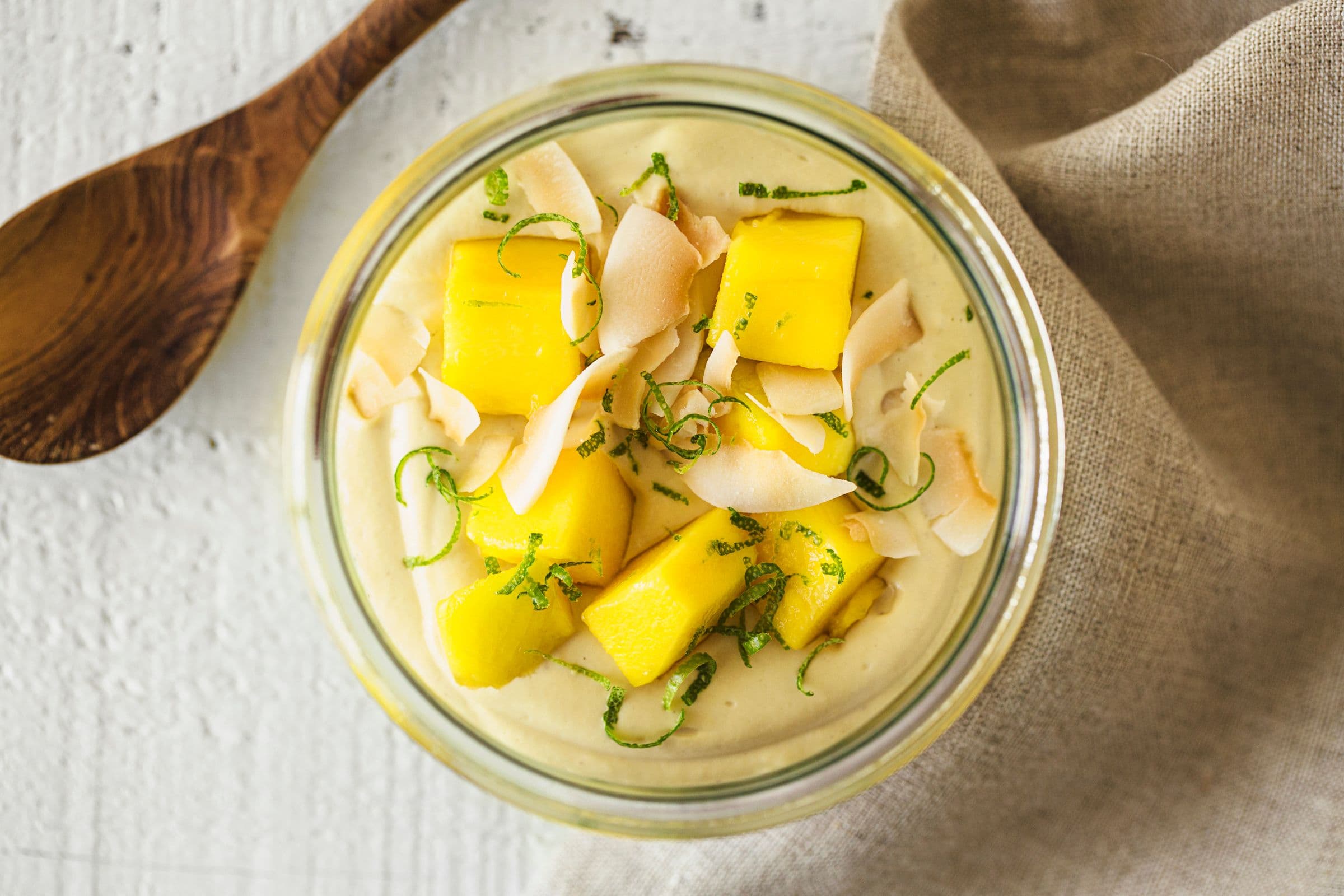Glass bowl of vegan mango cashew yogurt topped with fresh mango cubes, lime zest and toasted coconut flakes.