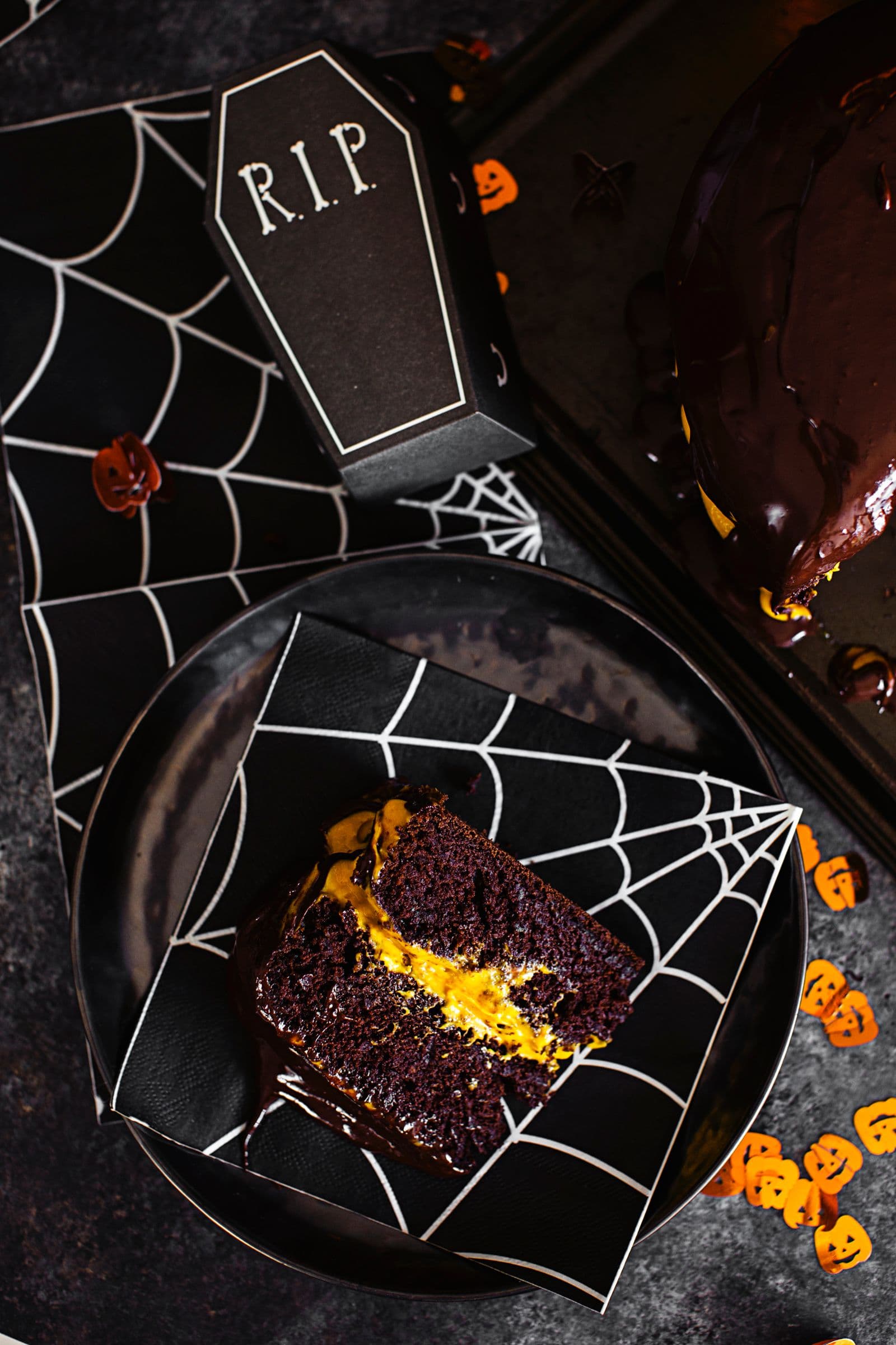 Porção de bolo vegan de Halloween servida num prato preto decorado com teias, com uma pequena caixa temática de caixão como elemento decorativo.
