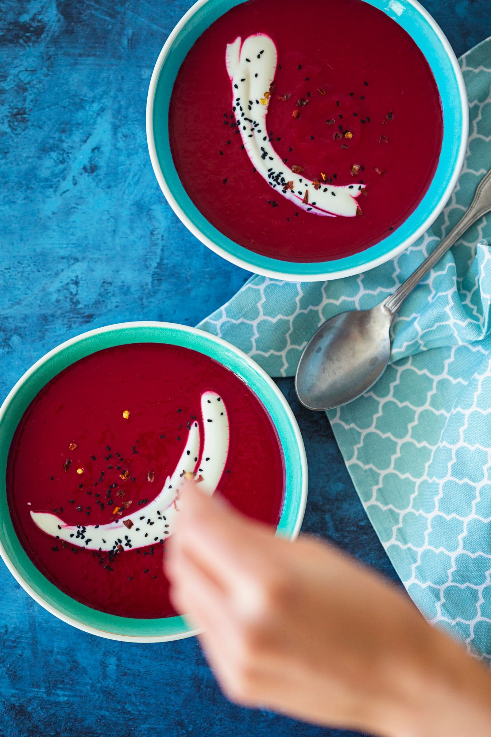 Hand drizzling a sprinkle of nigella seeds onto vibrant beetroot soup in a turquoise bowl.