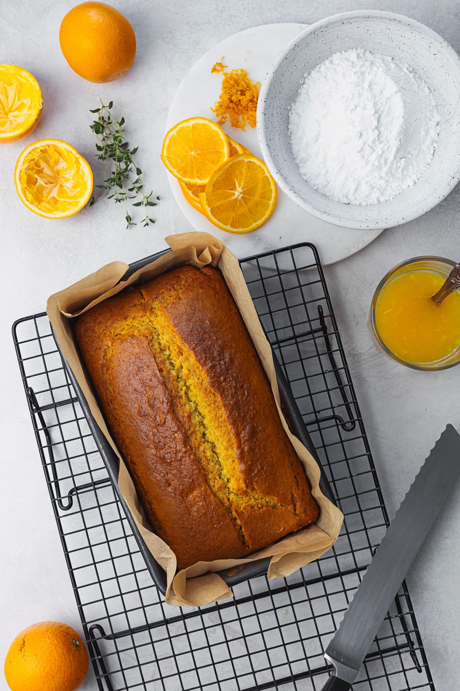 Bolo de laranja acabado de sair do forno, ainda na forma, sobre uma grelha de arrefecimento, com ingredientes da calda ao lado.