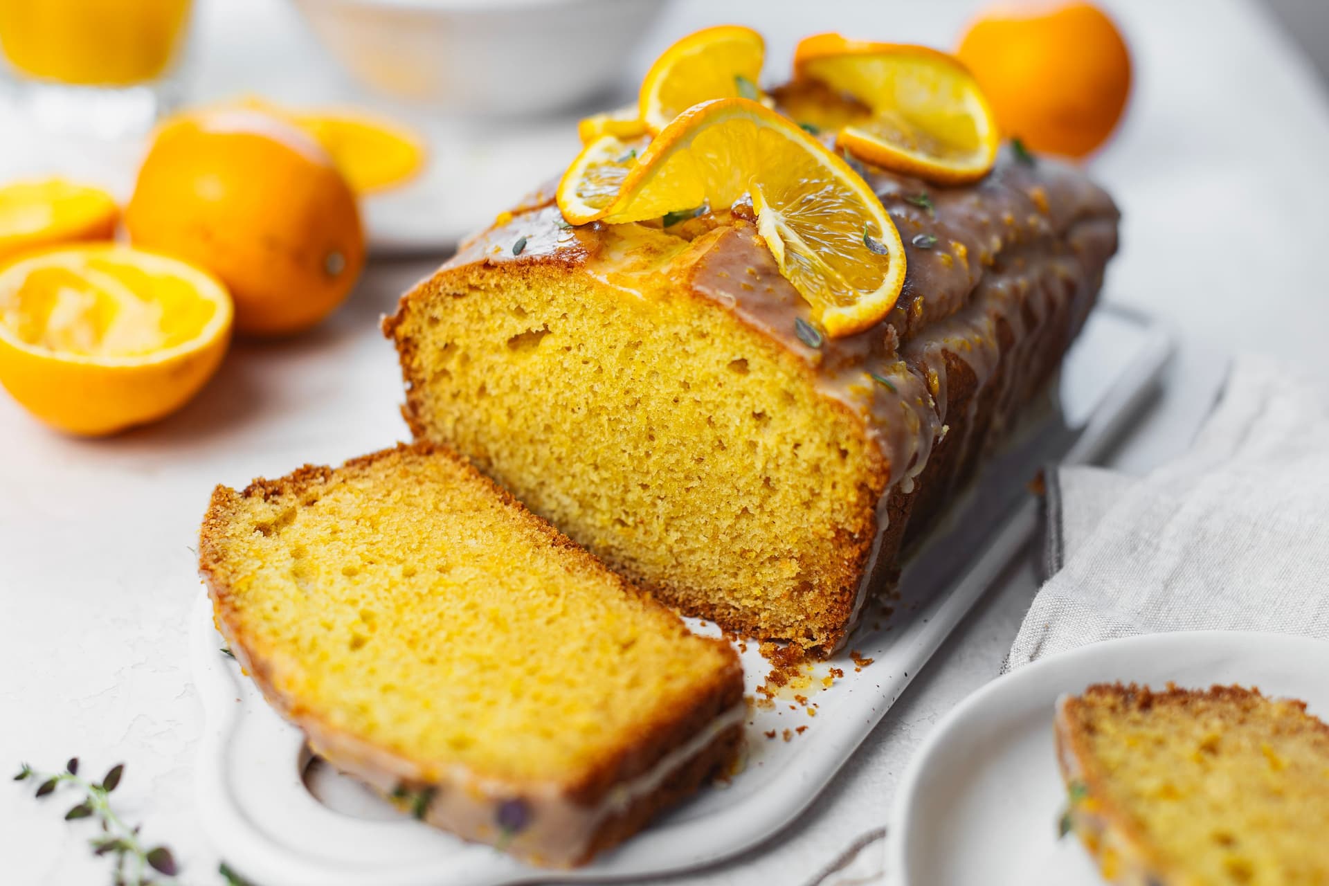Bolo de laranja fatiado com cobertura de calda de laranja brilhante, decorado com laranja fresca e tomilho, com laranjas ao fundo.