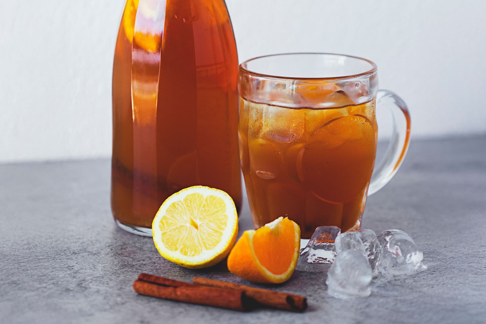 Copo e garrafa de chá gelado com rodelas de laranja e limão, paus de canela e cubos de gelo.