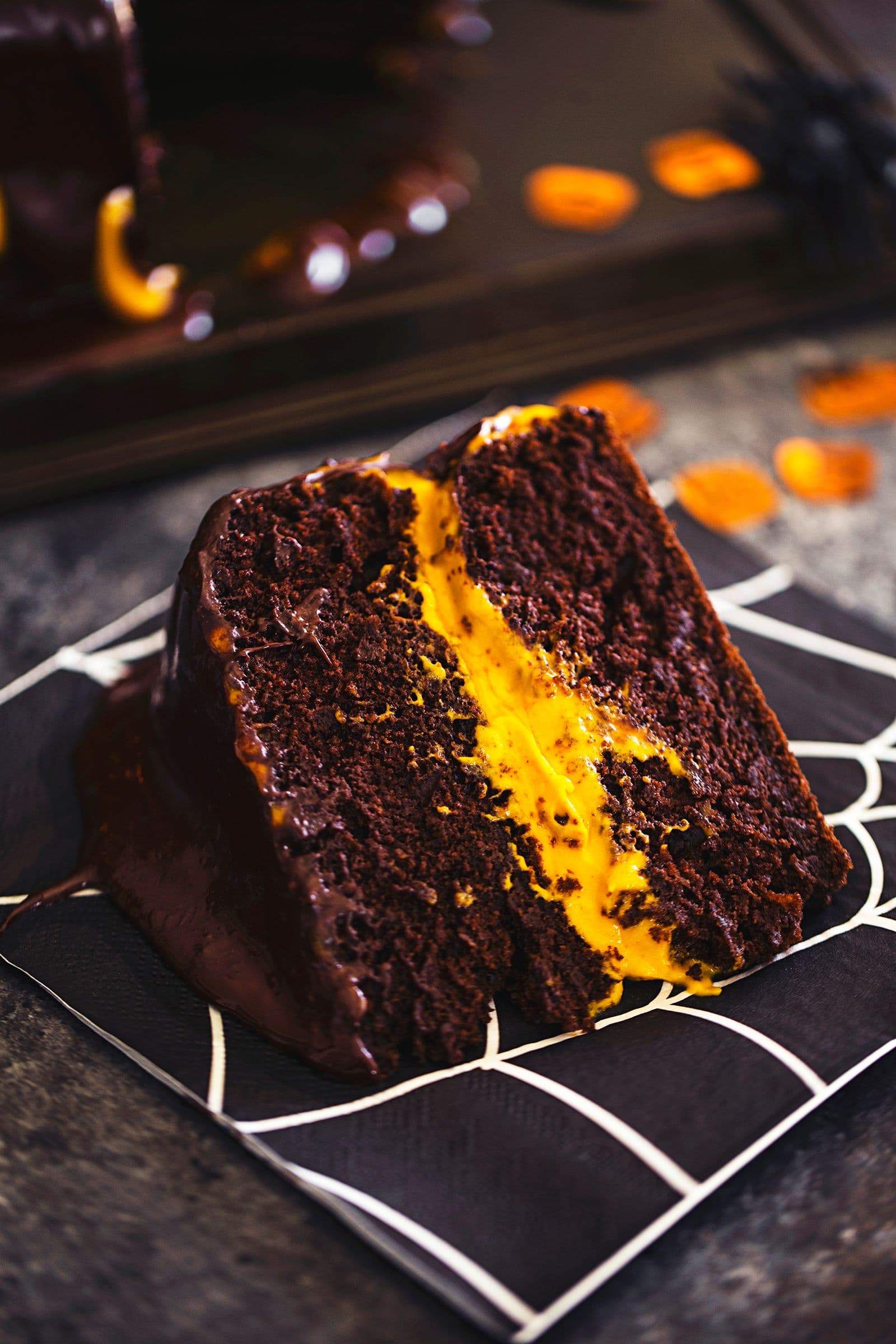 Slice of moist chocolate cake with bright orange cashew frosting, served on a black spiderweb-patterned napkin.