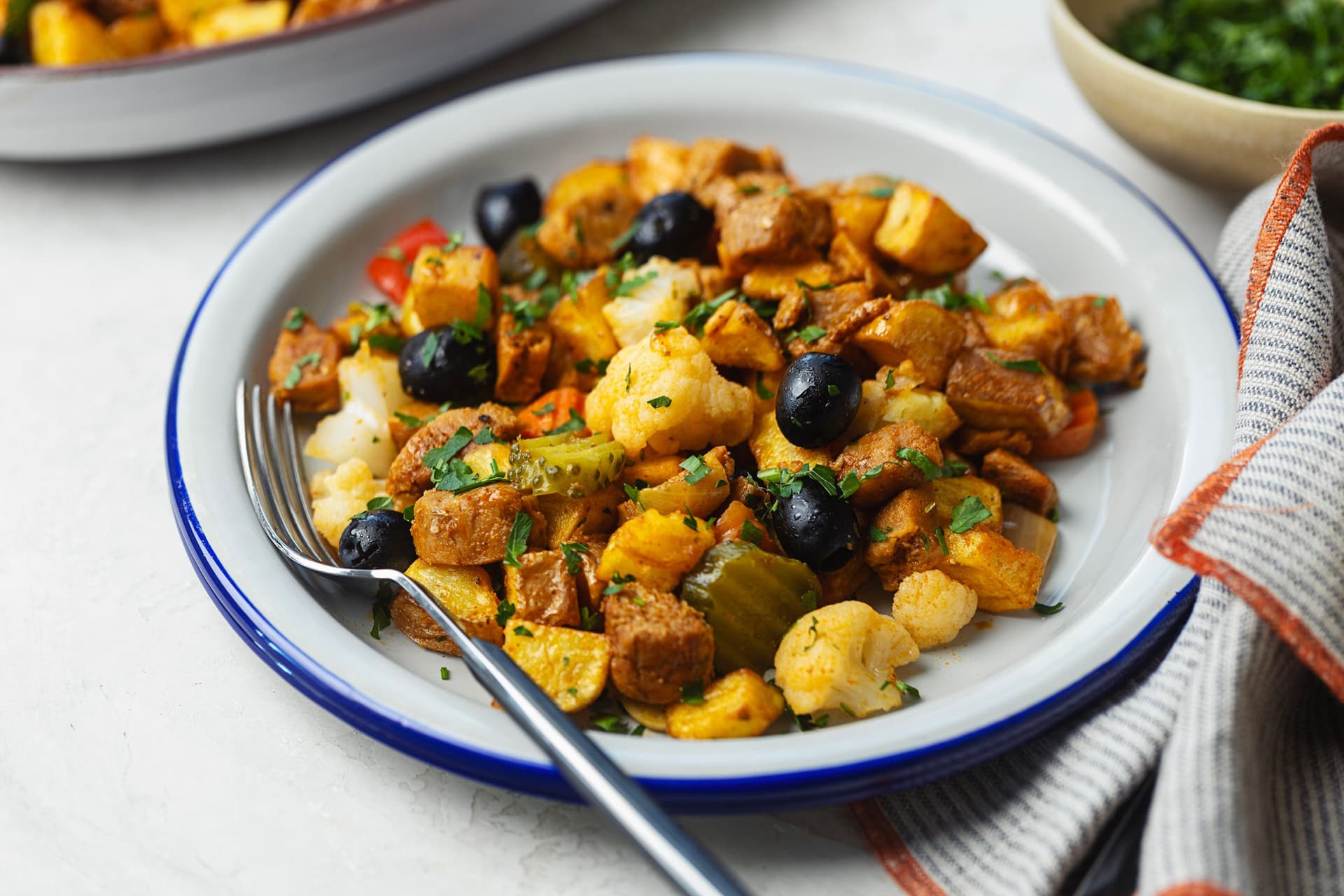 Prato de borda azul com porção de pica pau de soja fotografado em ângulo lateral, com azeitonas pretas, couve-flor, pickles, batatas e soja caramelizada com salsa fresca