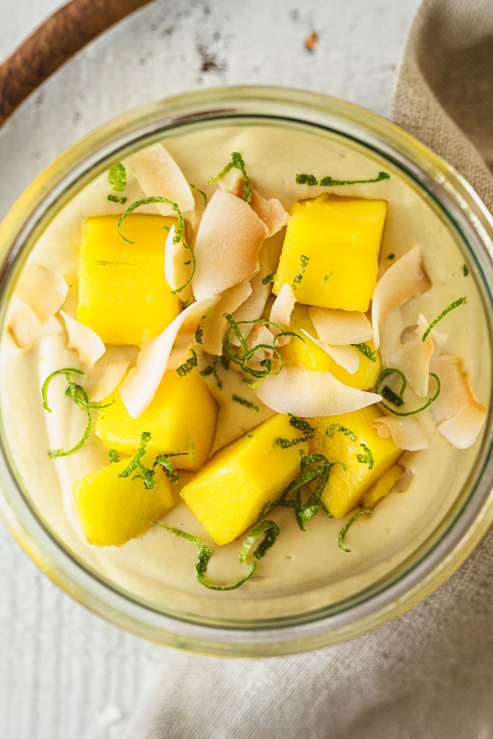 Glass bowl of vegan mango cashew yogurt topped with fresh mango cubes, lime zest and toasted coconut flakes.