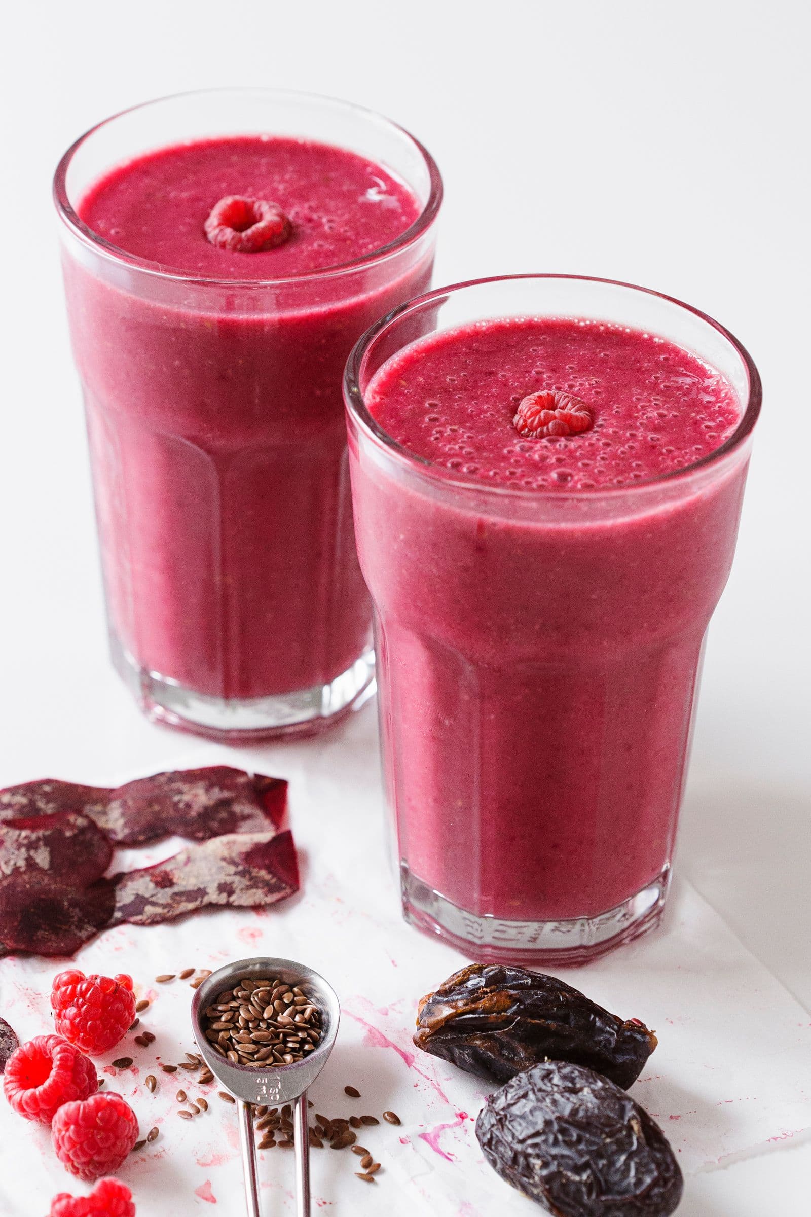 Two glasses of beet raspberry smoothie on a white surface with scattered dates, berries and flax seeds.