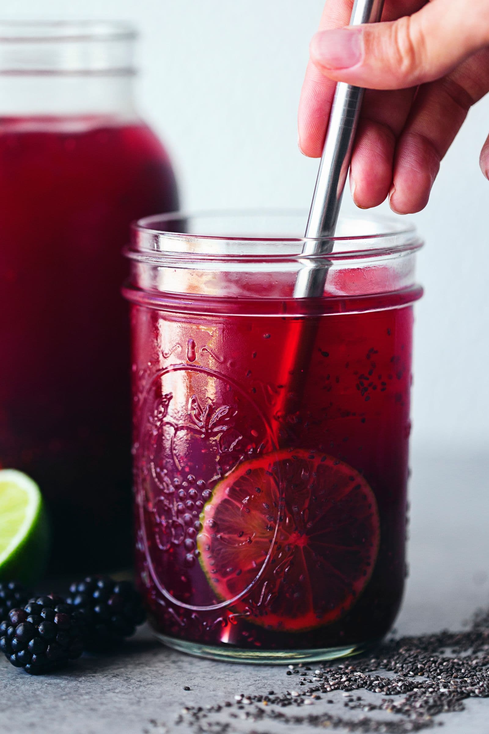 Mão a mexer um copo de chia fresca com lima e amoras em segundo plano.