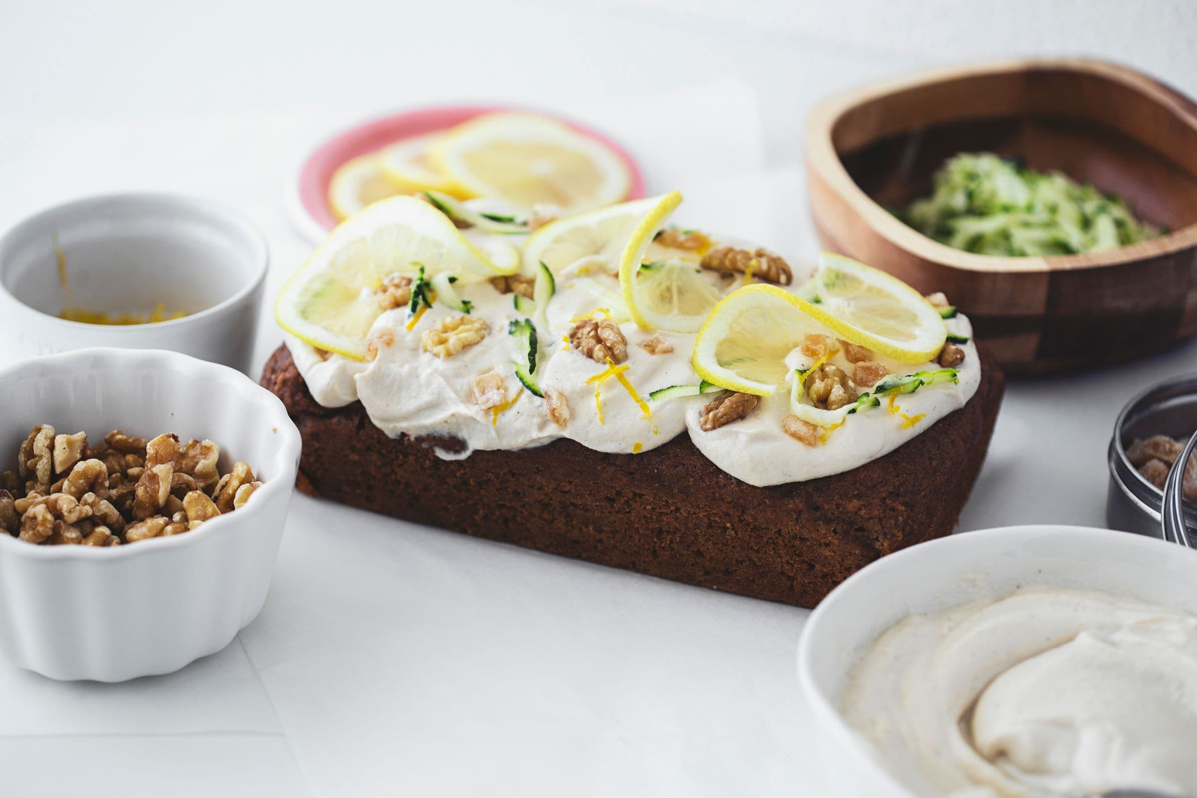 Zucchini and lemon cake on a white table with bowls of walnuts and frosting nearby.