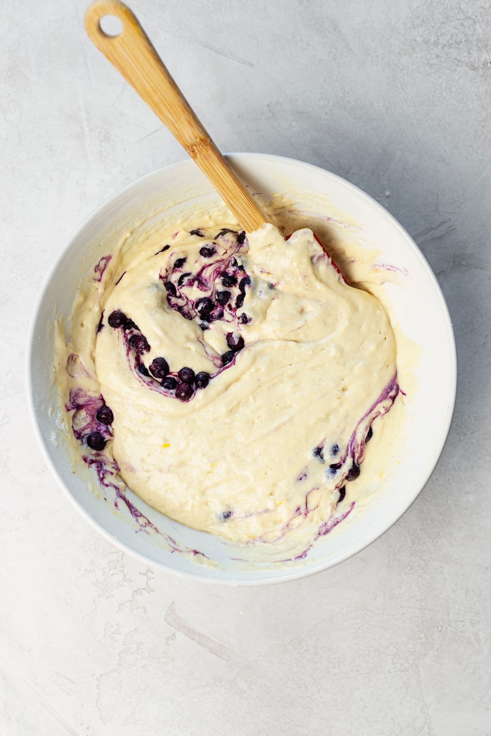 Muffin batter with blueberries and lemon zest being gently folded in a mixing bowl.