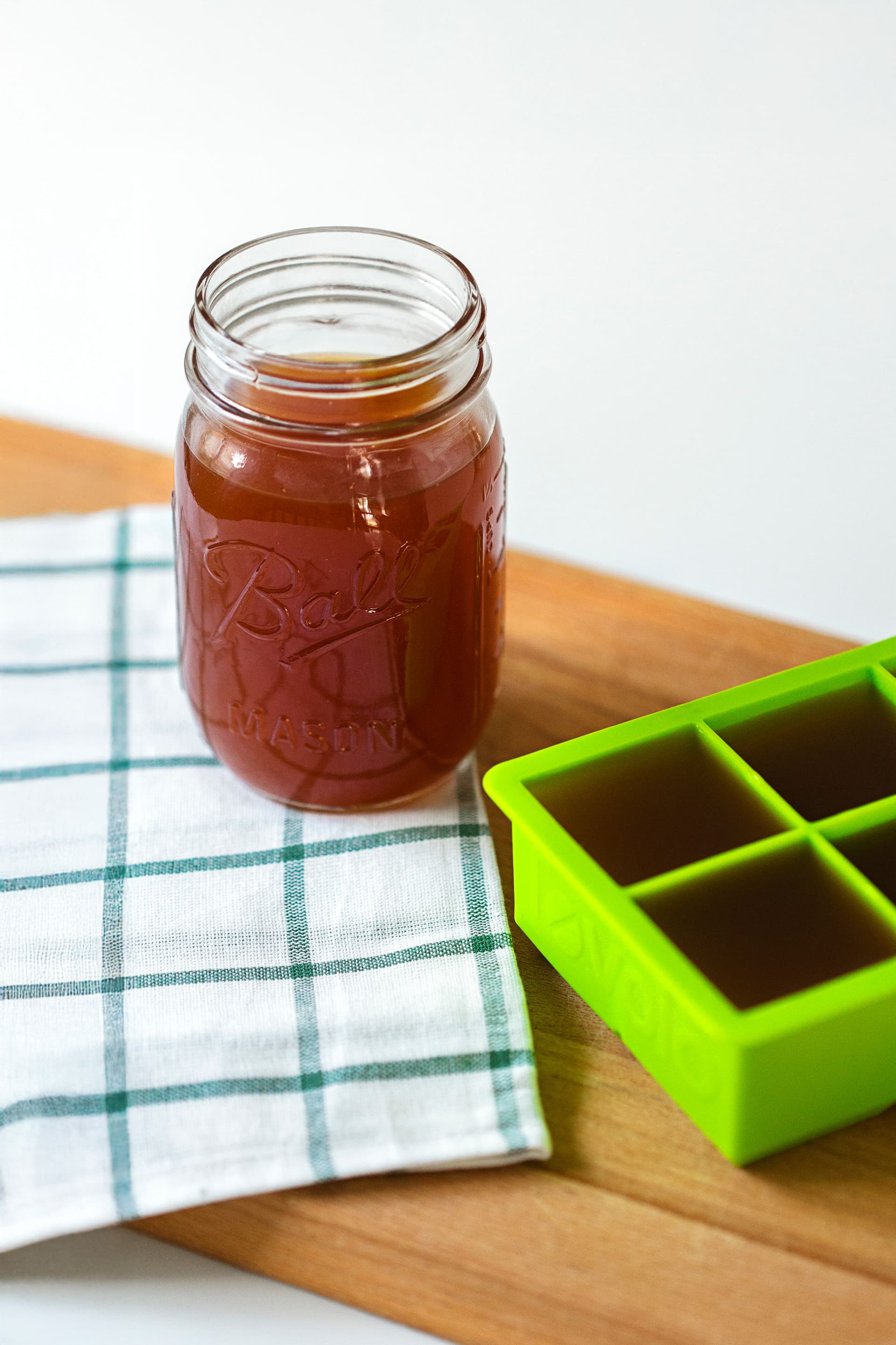 Frasco de vidro com caldo de legumes caseiro ao lado de um tabuleiro verde para congelação em cubos.