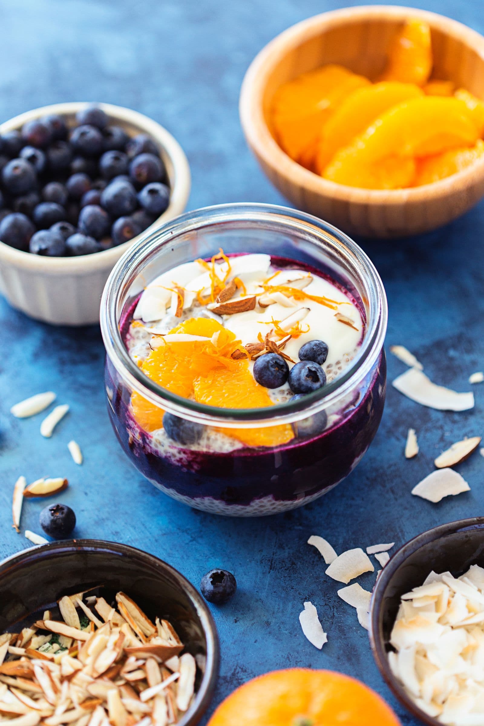 Frasco de pudim de chia rodeado por taças com mirtilos, gomos de laranja e amêndoas picadas.