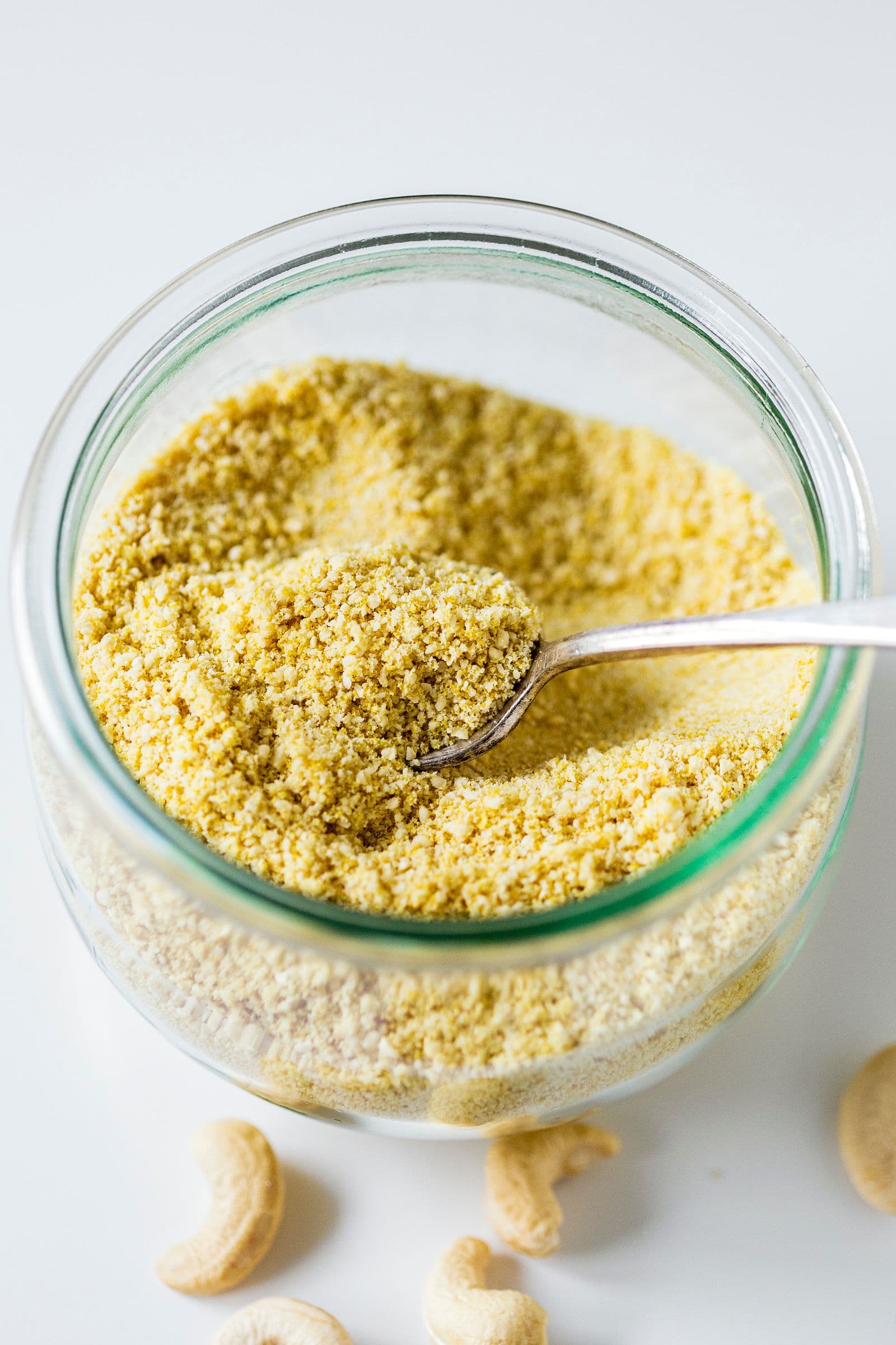 Glass jar with vegan parmesan and a spoon, surrounded by cashews.