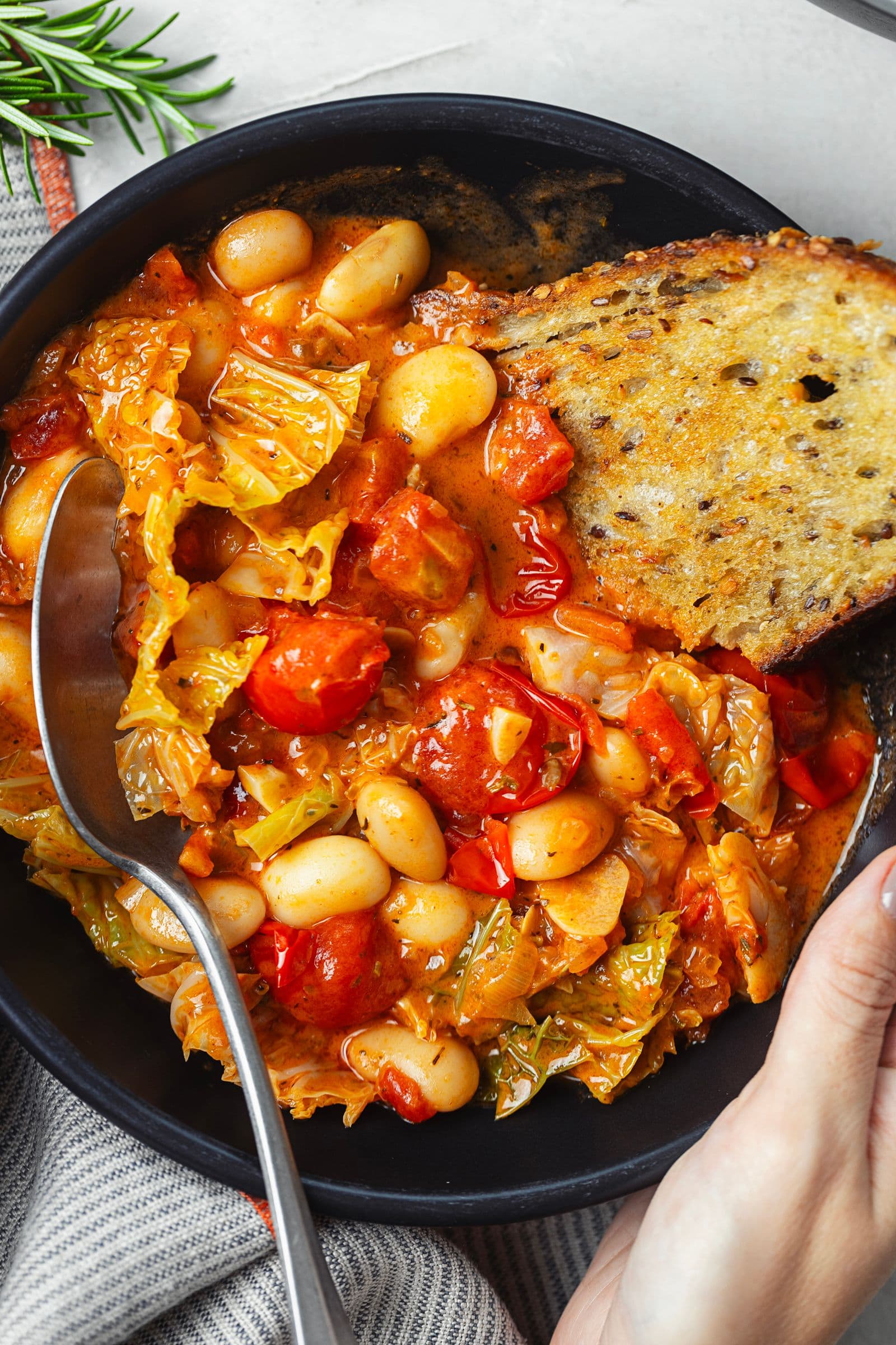 Close-up of the stew in a black bowl, showing the beans, cherry tomatoes, cabbage and a slice of toasted bread.