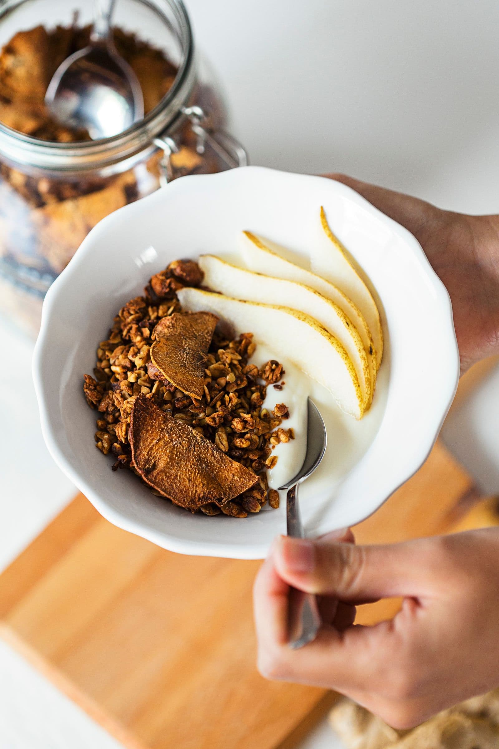 Mãos a segurar uma taça de granola e iogurte sobre uma tábua de madeira.