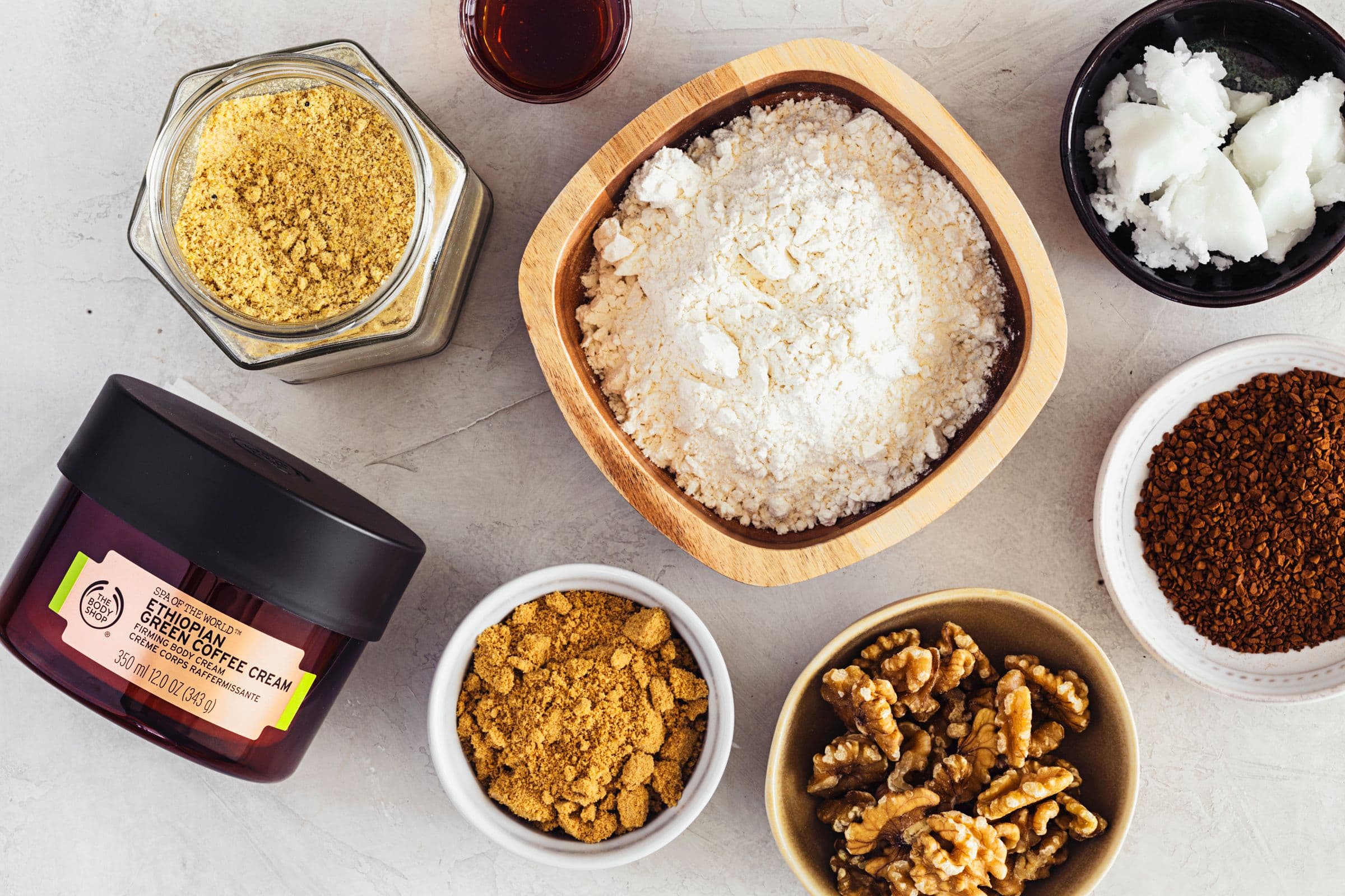 Vista de cima dos ingredientes para o bolo de café e pêra: farinha, açúcar, café, óleo de coco, nozes, e um frasco de creme de Café Verde da Etiópia da The Body Shop