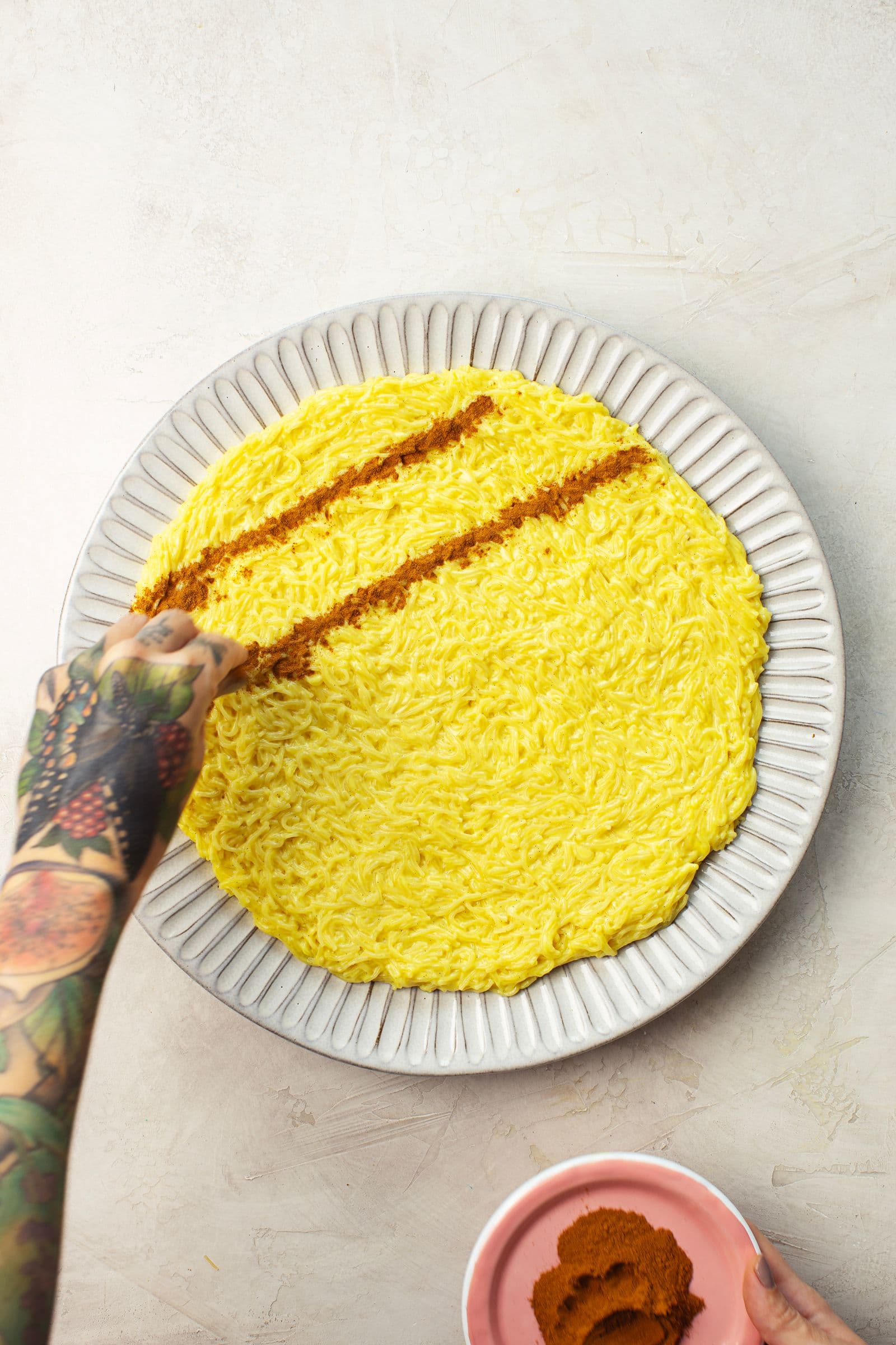 Overhead view of vegan Aletria being decorated with cinnamon, with a tattooed hand creating diagonal lines across the surface.