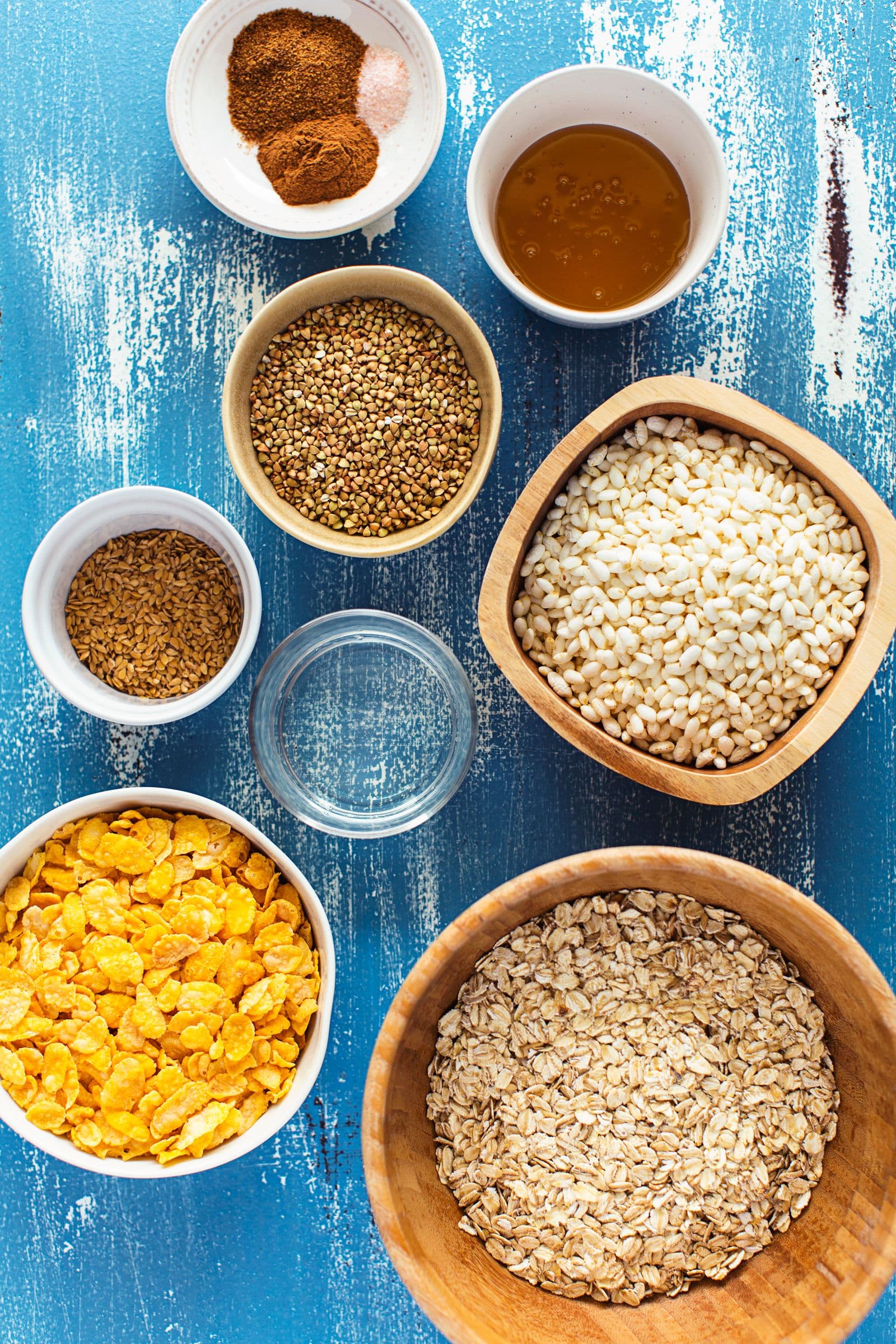 Vista de cima dos ingredientes para a granola em taças: flocos de aveia, cornflakes, sementes e especiarias.