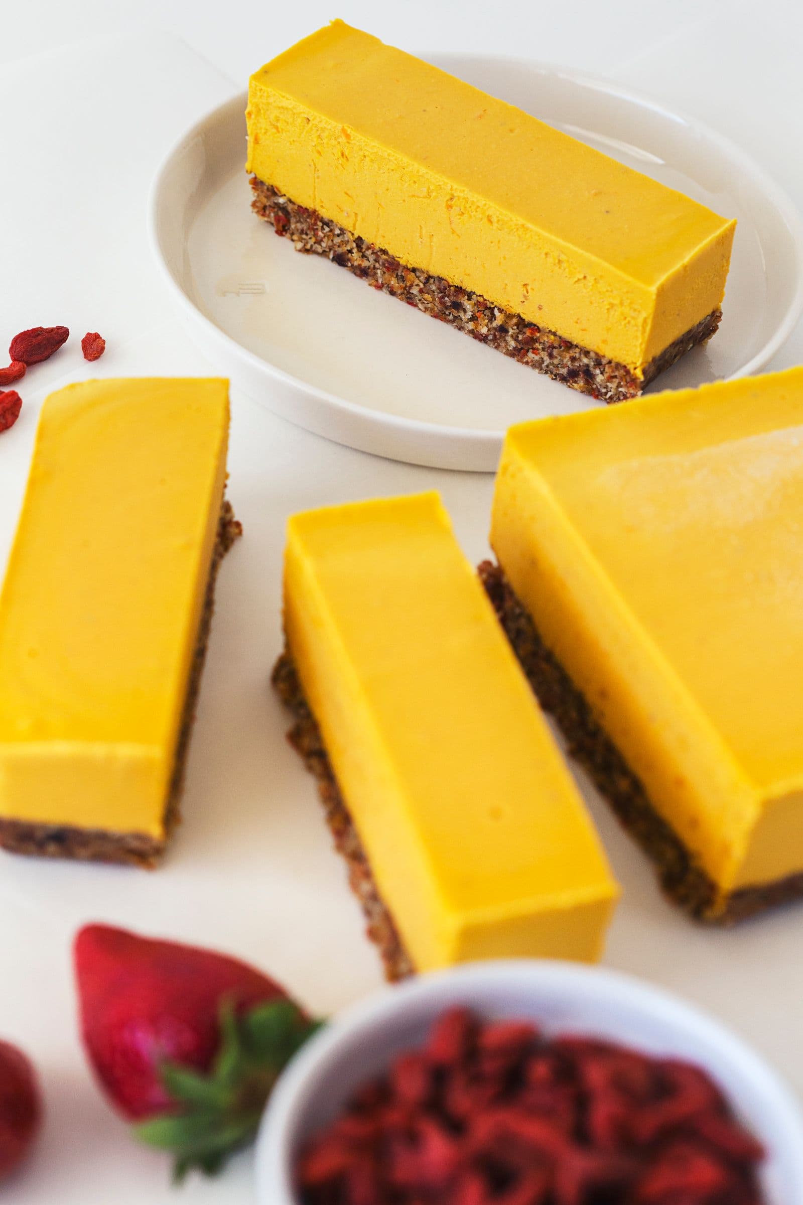Sliced cheesecake bars with bowls of goji berries and strawberries nearby.