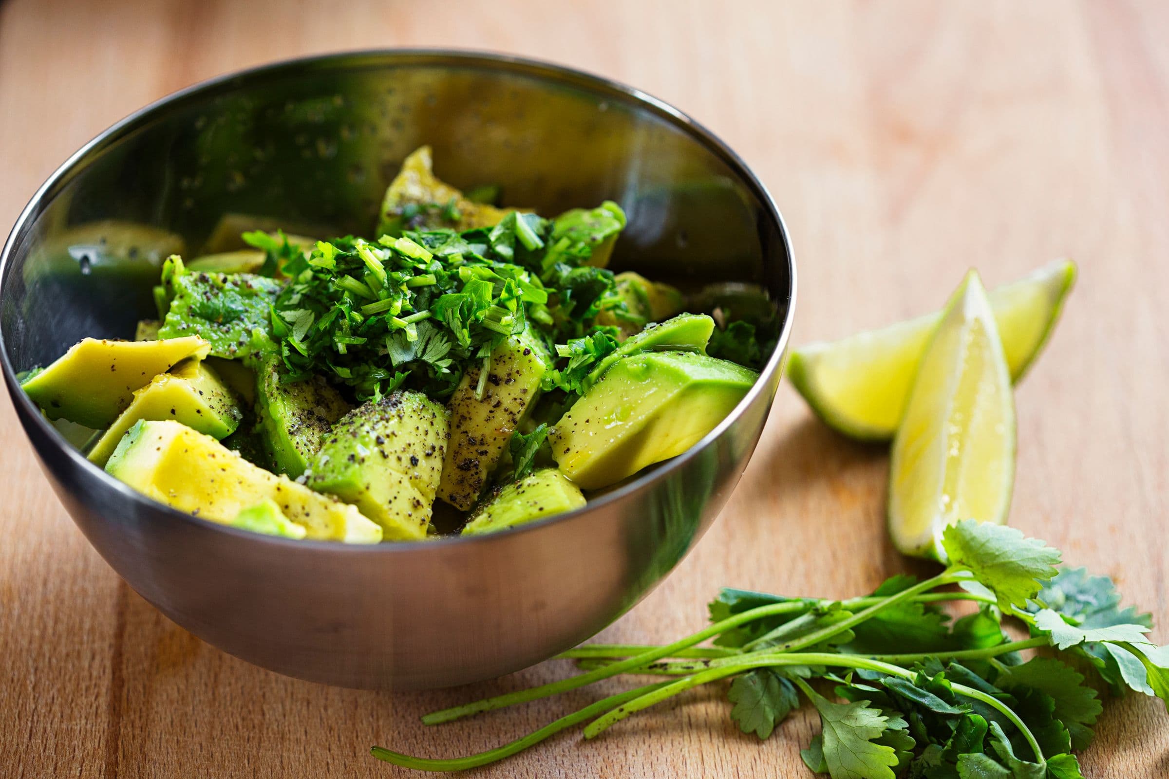 Metal bowl with avocado chunks, coriander and spices on a wooden surface with lime wedges.