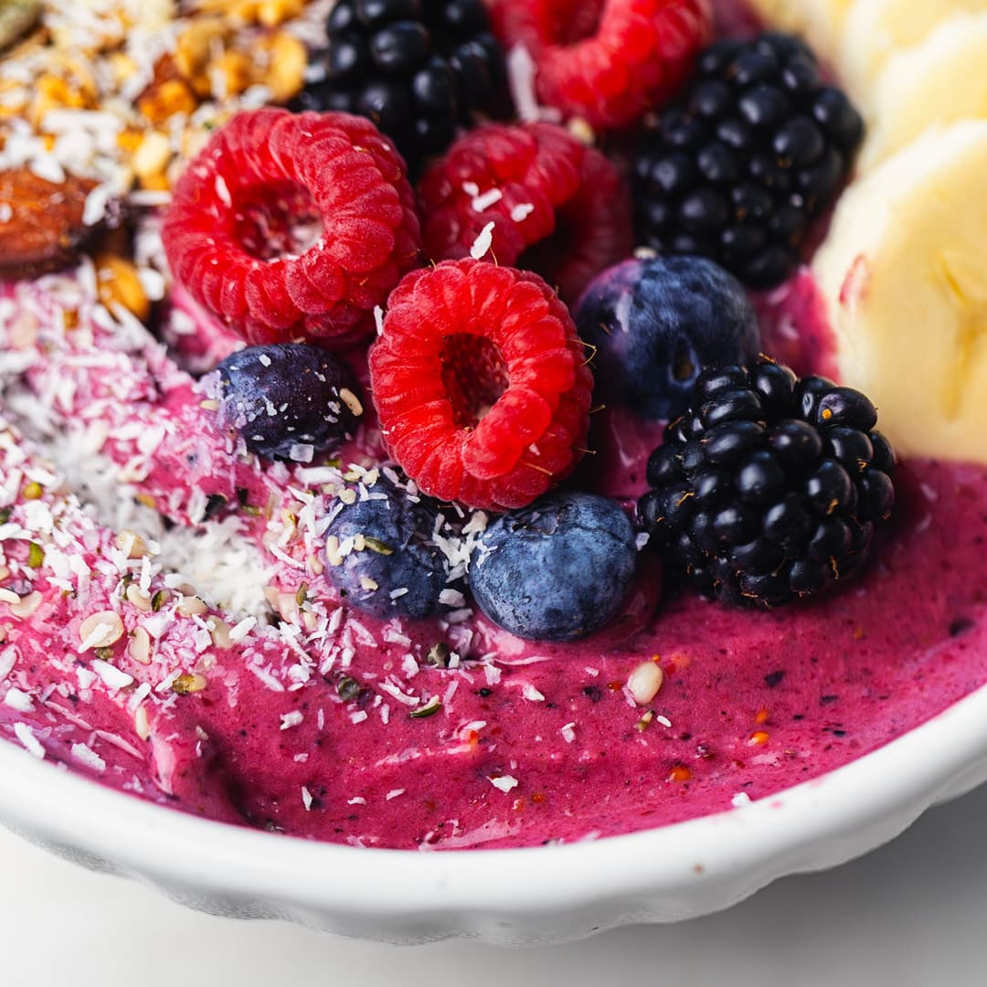 Taça de smoothie de frutos vermelhos com banana, framboesas, amoras, mirtilos, coco e granola.