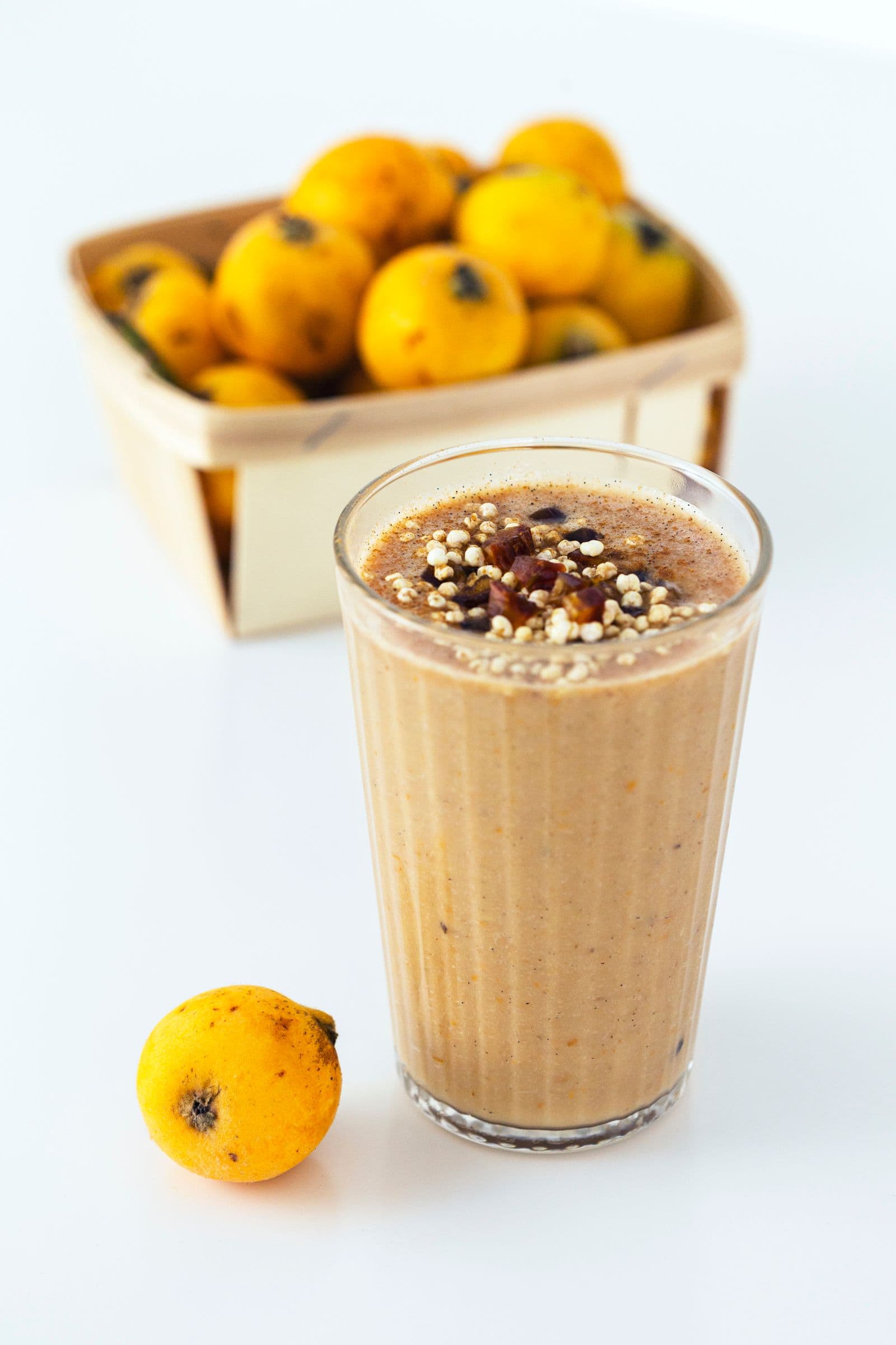 Loquat smoothie in a glass topped with puffed millet and chopped medjoul date, with a fresh fruit basket and one loquat in the foreground