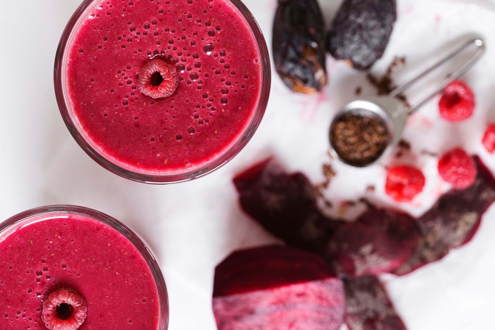 Dois smoothies cor-de-rosa vibrante com beterraba e framboesa, com frutas, tâmaras e sementes de linhaça espalhadas à volta.