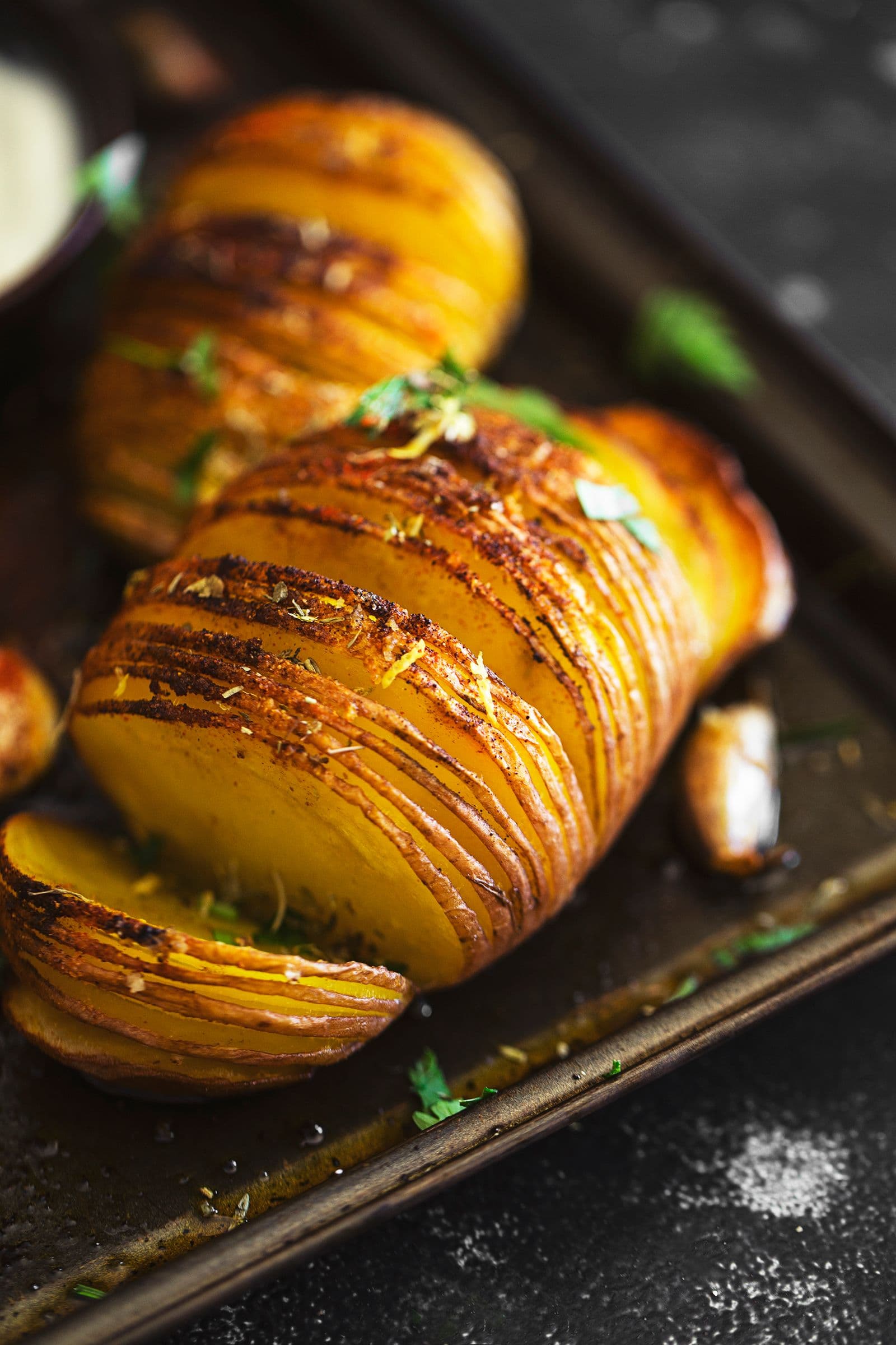 Close view of a seasoned Hasselback potato on a dark baking tray with roasted garlic.